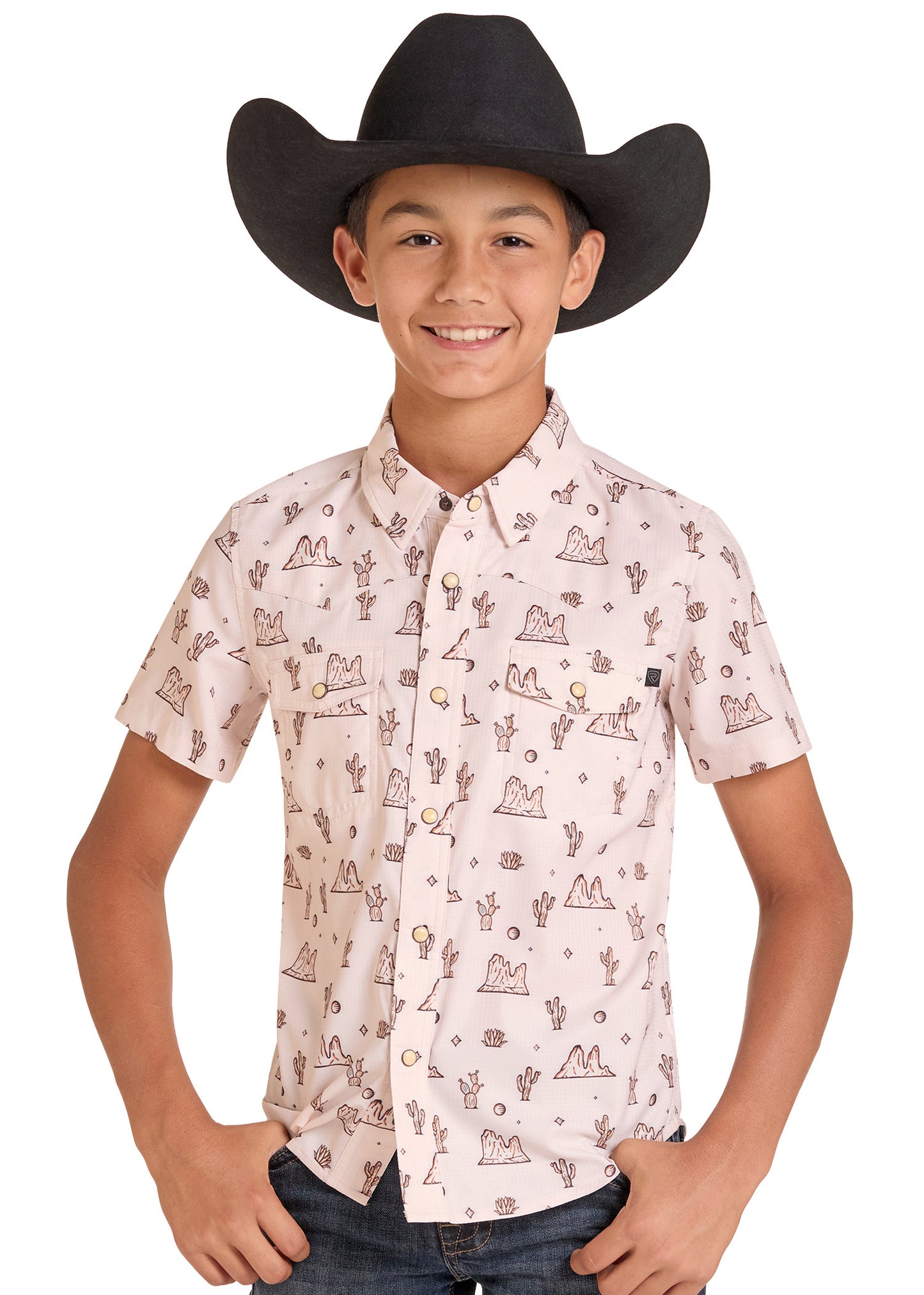 A young boy wearing a black cowboy hat and a light pink short-sleeve button-up shirt with a brown western-themed print of cacti, mountains, and coins, paired with dark jeans against a white background.