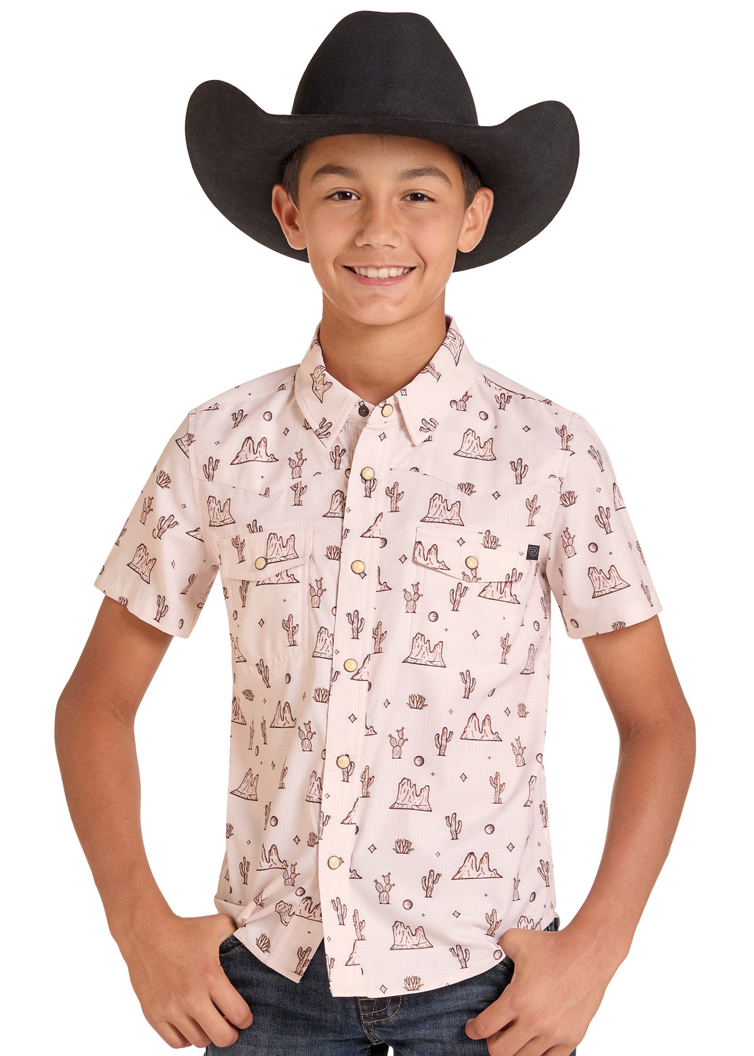 A young boy wearing a black cowboy hat and a light pink short-sleeve button-up shirt with a brown western-themed print of cacti, mountains, and coins, paired with dark jeans against a white background.
