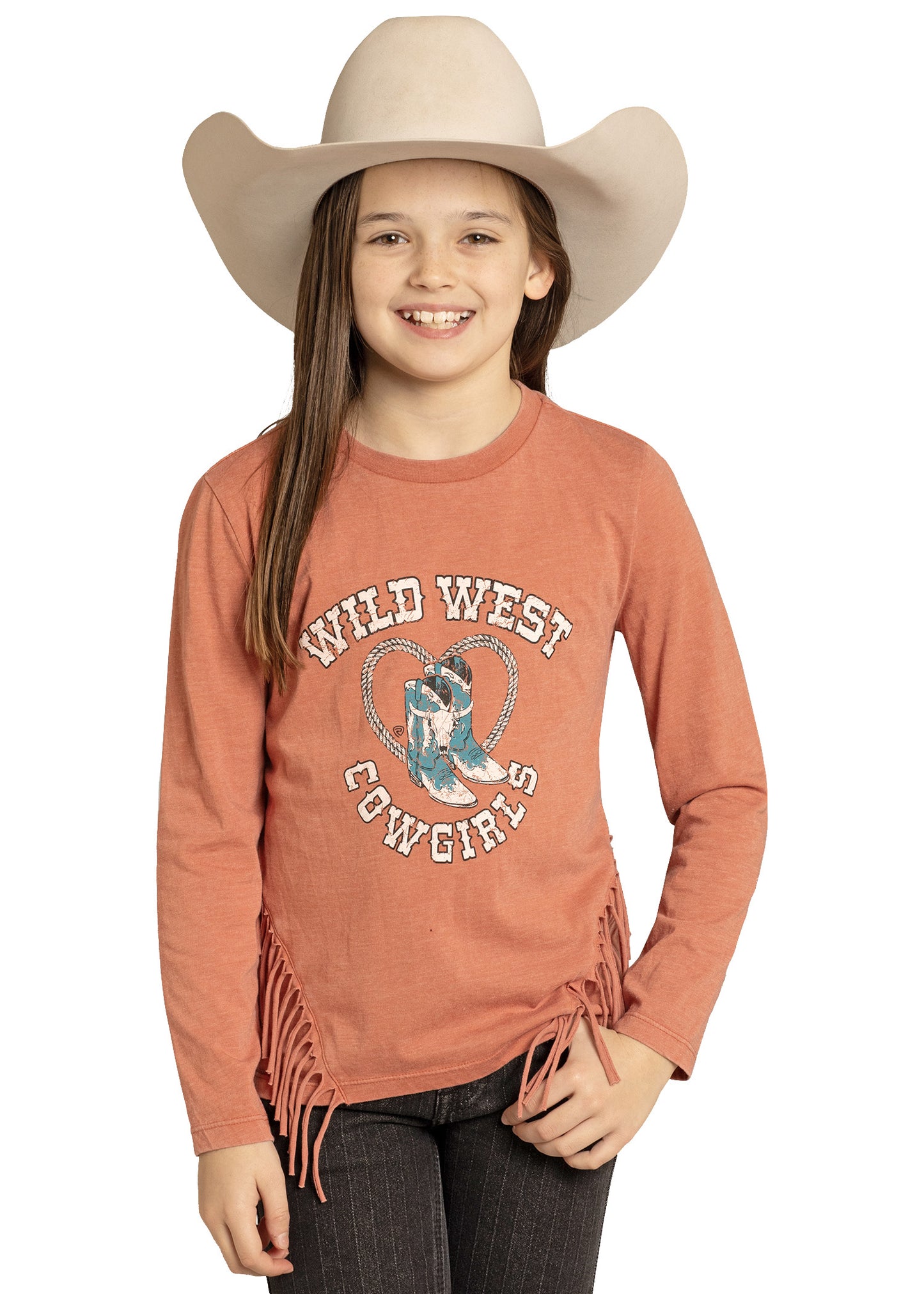 A smiling girl wears the Rock & Roll Denim Girls' Wild West Fringe Top, a rust-colored long sleeve tee with fringe, “Wild West Cowgirls” text, and cowboy boots graphic, adding classic Western charm against a white background.