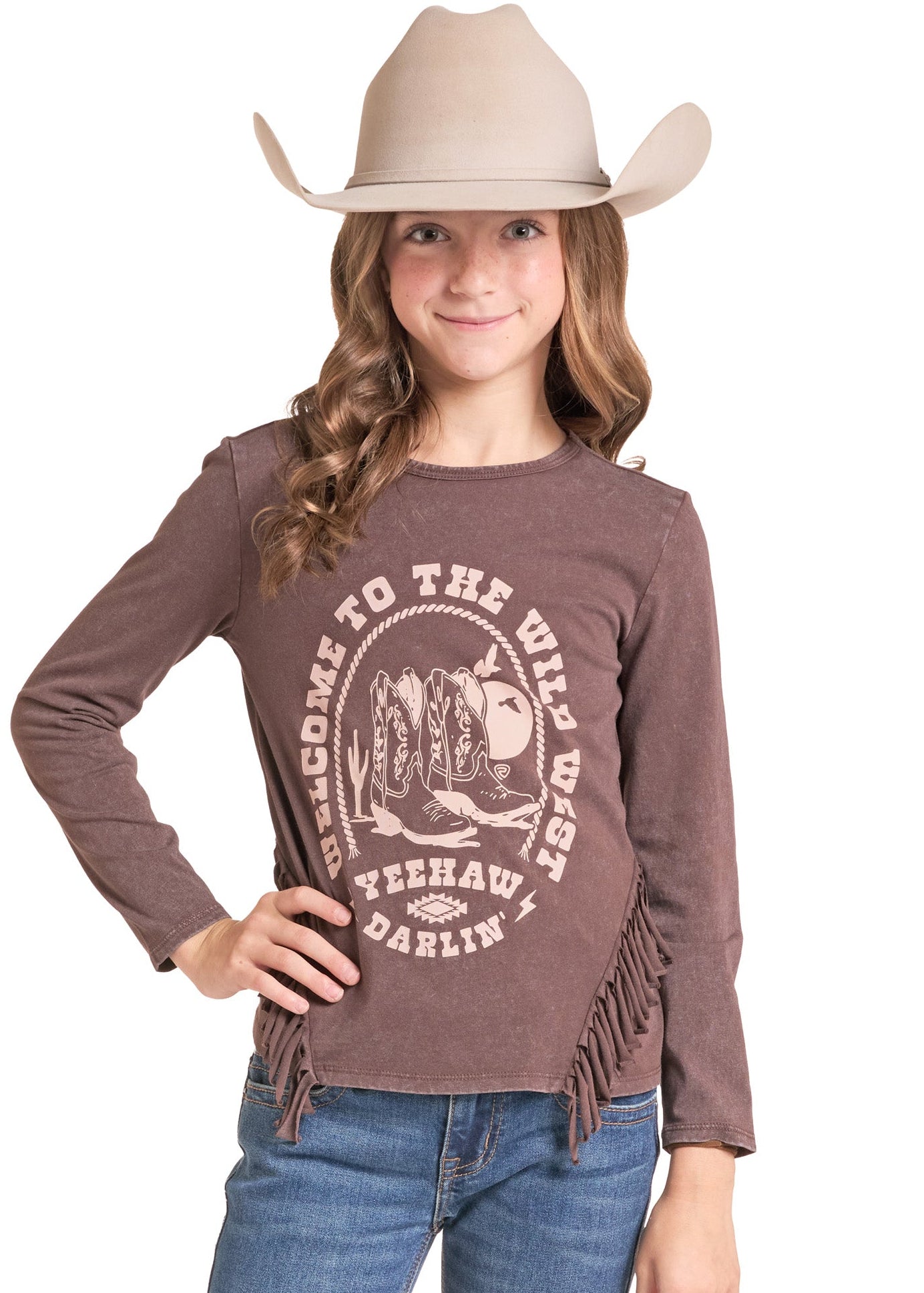 A young girl wearing a brown long-sleeve t-shirt with fringe and a western graphic print, paired with blue jeans and a beige cowboy hat, against a plain white background.