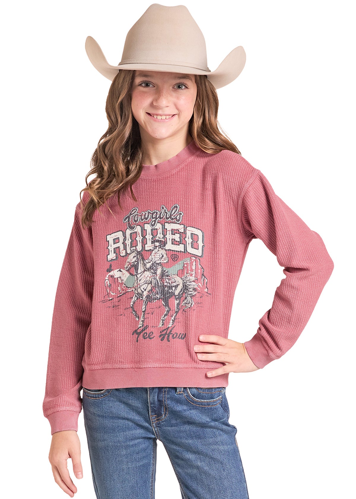 A young girl wearing a dusty rose waffle-knit sweatshirt with a western graphic that reads 'Tough Rodeo', paired with blue jeans and a beige cowboy hat.