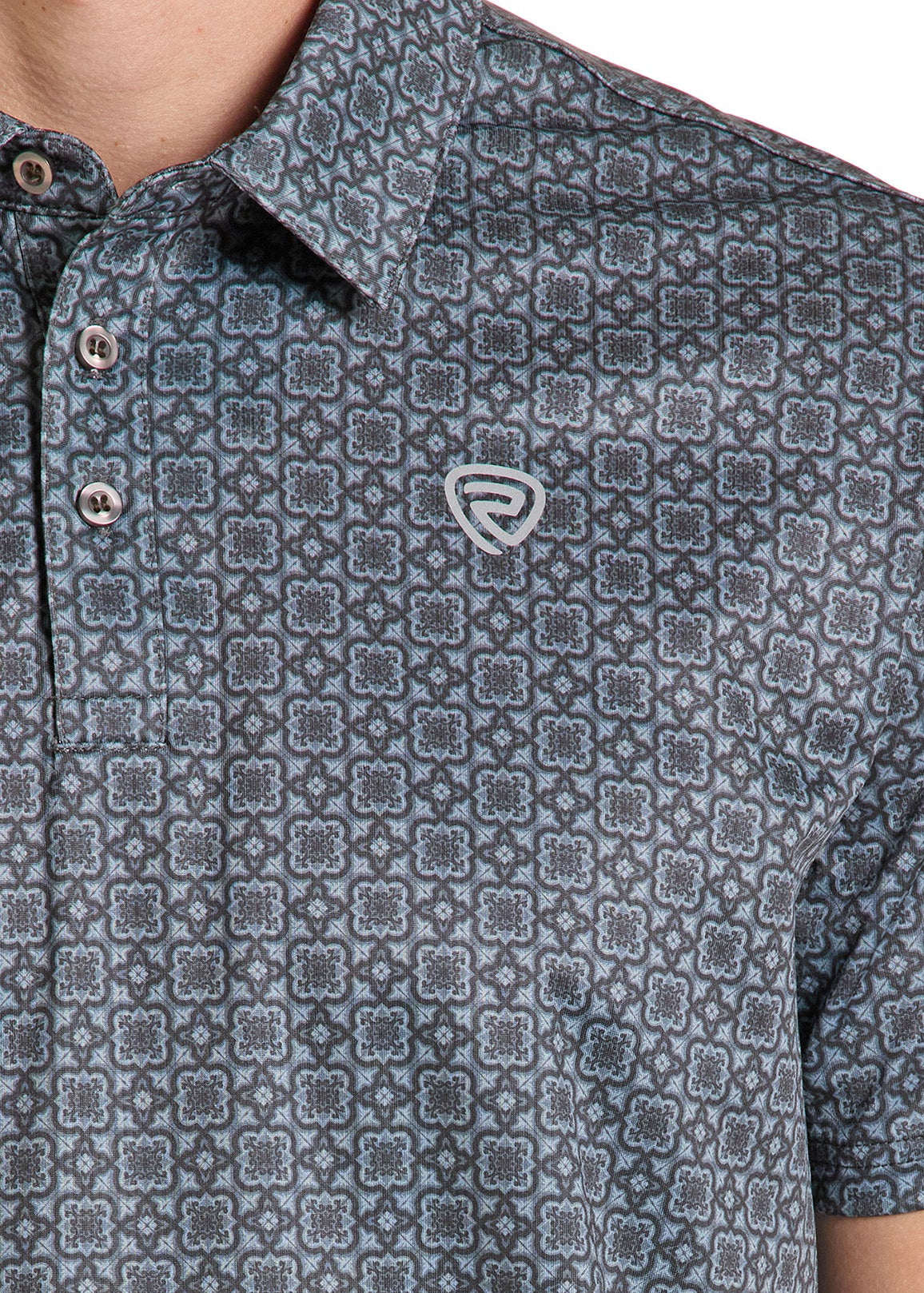 Close-up of a man wearing a short-sleeve polo shirt with a repeating geometric pattern in dark gray and blue, featuring a small embroidered shield logo on the chest.