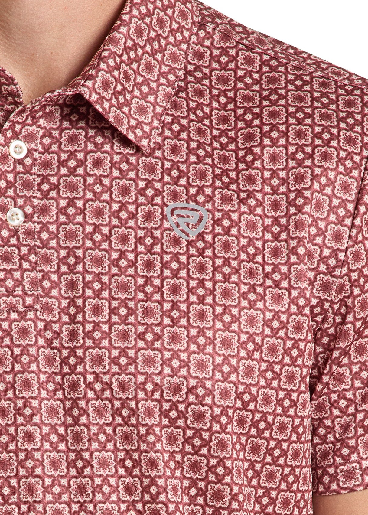 Close-up of a short-sleeve collared polo shirt with an allover geometric pattern in dusty rose and white, featuring a small embroidered chest logo.