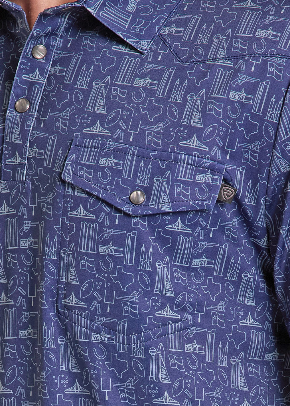 Close-up of a man wearing a short-sleeve button-up shirt in blue with a white line-art pattern of boats, ships, and maritime icons. The shirt features a chest pocket with a snap button and a small circular brand patch.
