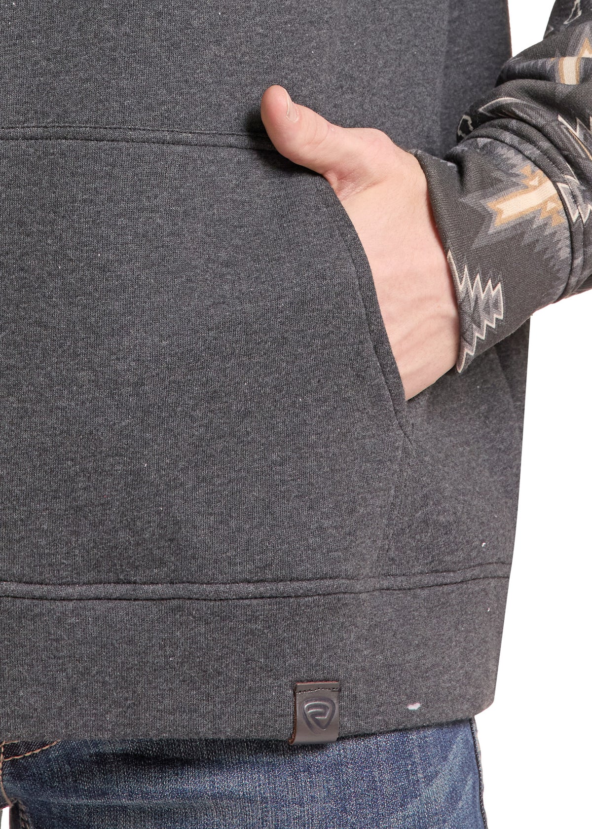 Close-up of a person wearing a dark heather gray pullover hoodie with a kangaroo pocket, their hand in the pocket, and a patterned long-sleeve shirt visible underneath.