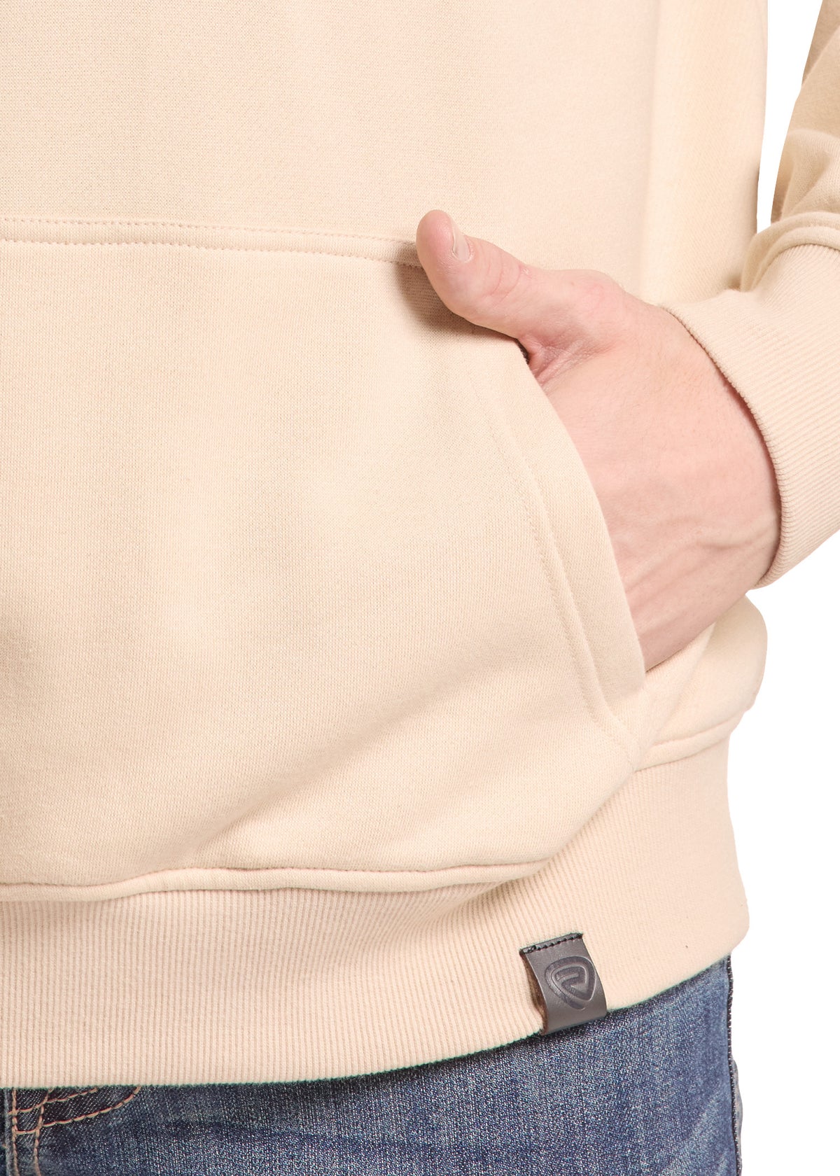 Close-up of a person wearing a light beige hoodie with a front pocket, hand in pocket, and blue jeans against a white background.