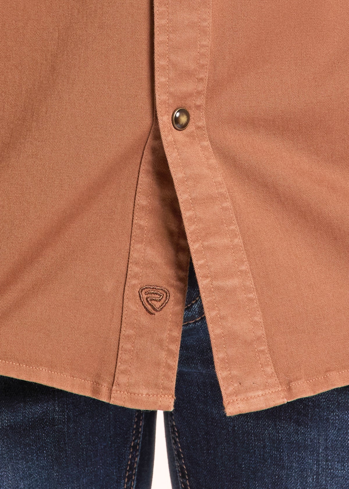 Close-up of a terracotta brown button-down shirt worn with dark blue jeans, showing the button closure and a small embroidered logo on the pocket.