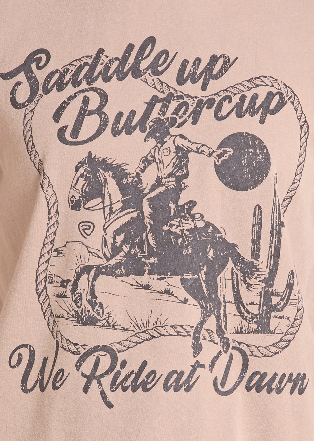 A close-up of a light beige graphic t-shirt featuring a western design with a cowboy riding a horse, a cactus, and the text 'Saddle up Buttercup' and 'We Ride at Dawn' in distressed black lettering.