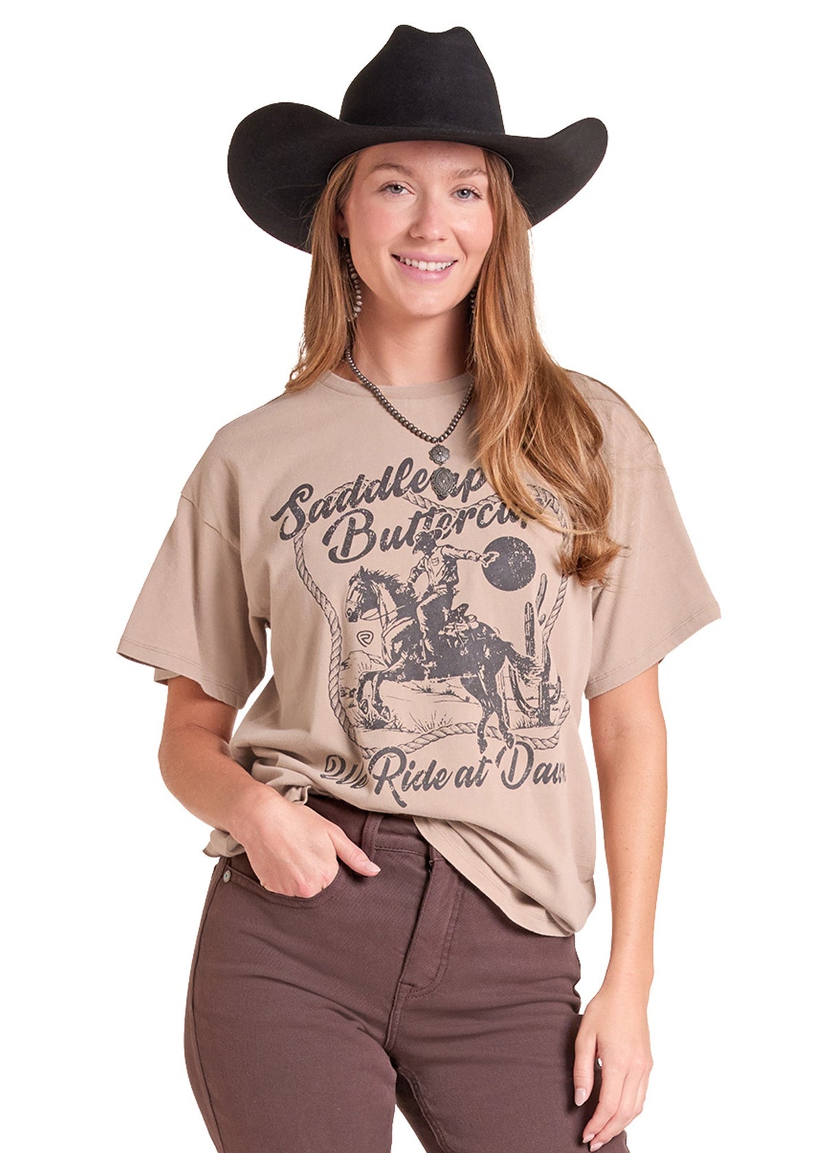 A woman modeling a beige graphic t-shirt with a western design that reads 'Saddle Up Butterfield Ride at One', paired with brown pants and a black cowboy hat, against a plain white background.