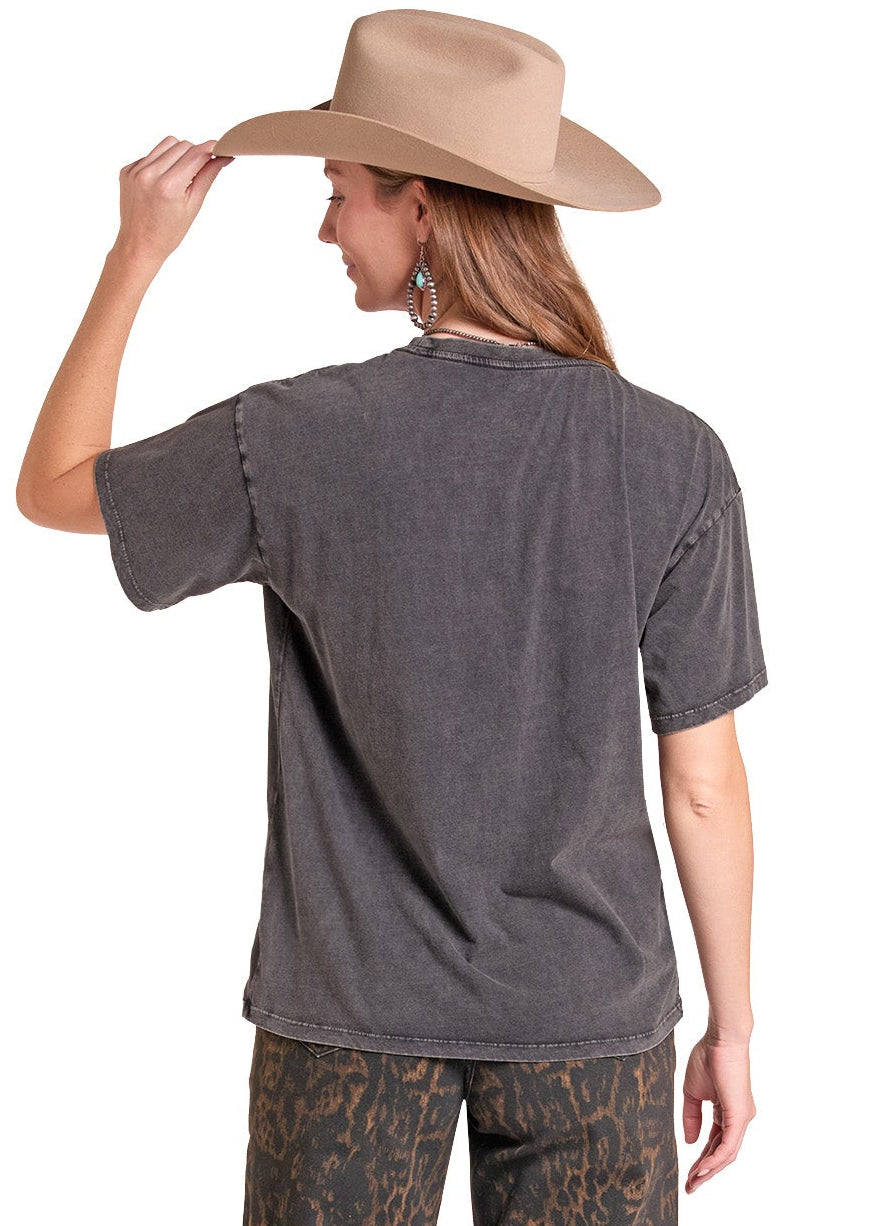 A woman shown from the back wearing a faded charcoal gray t-shirt, brown and black patterned wide-leg pants, a beige wide-brim hat, and white cowboy boots against a plain white background.