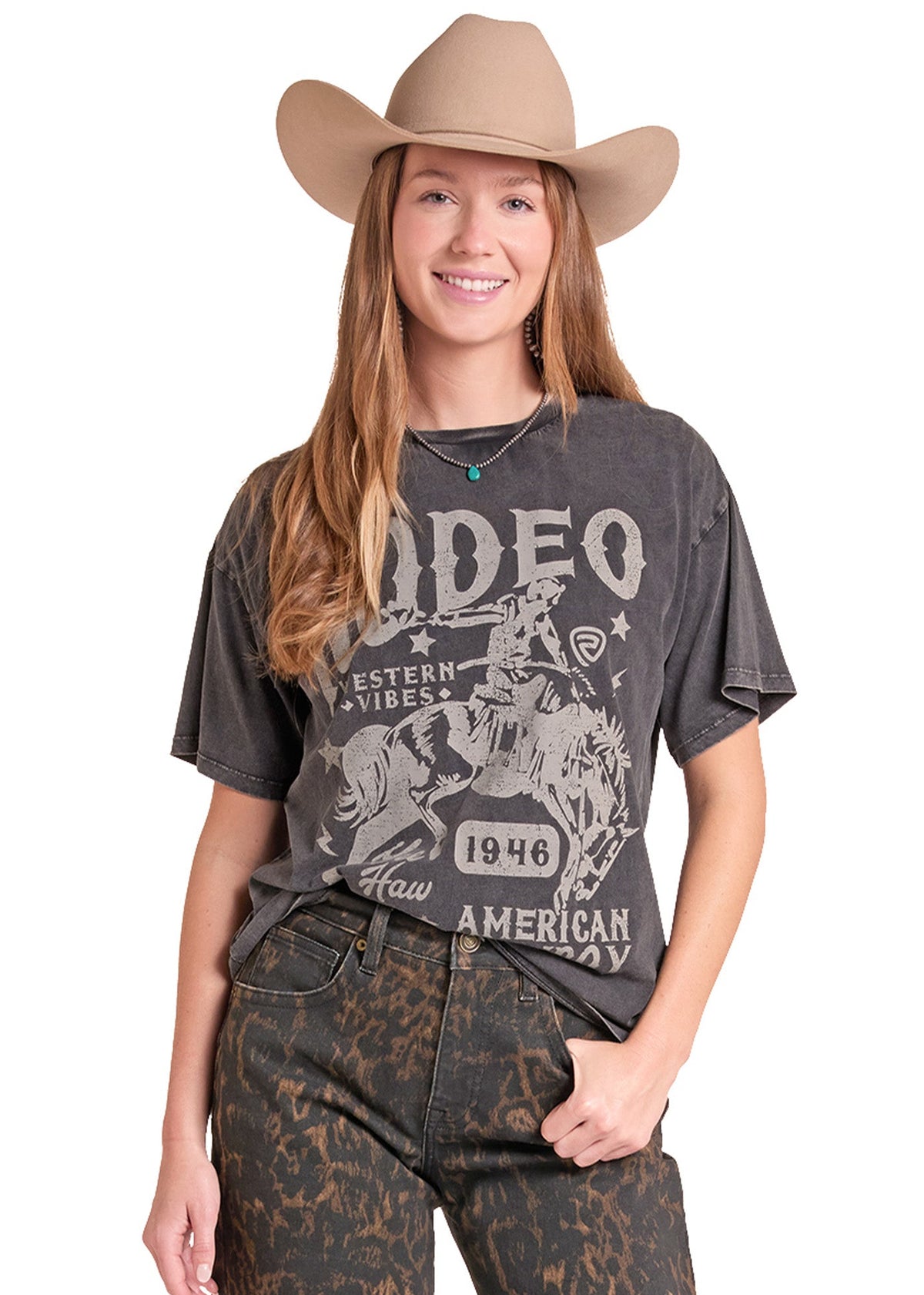 A woman modeling a dark gray acid-wash graphic t-shirt with a western 'Rodeo' design, paired with brown patterned jeans and a tan cowboy hat, against a plain white background.