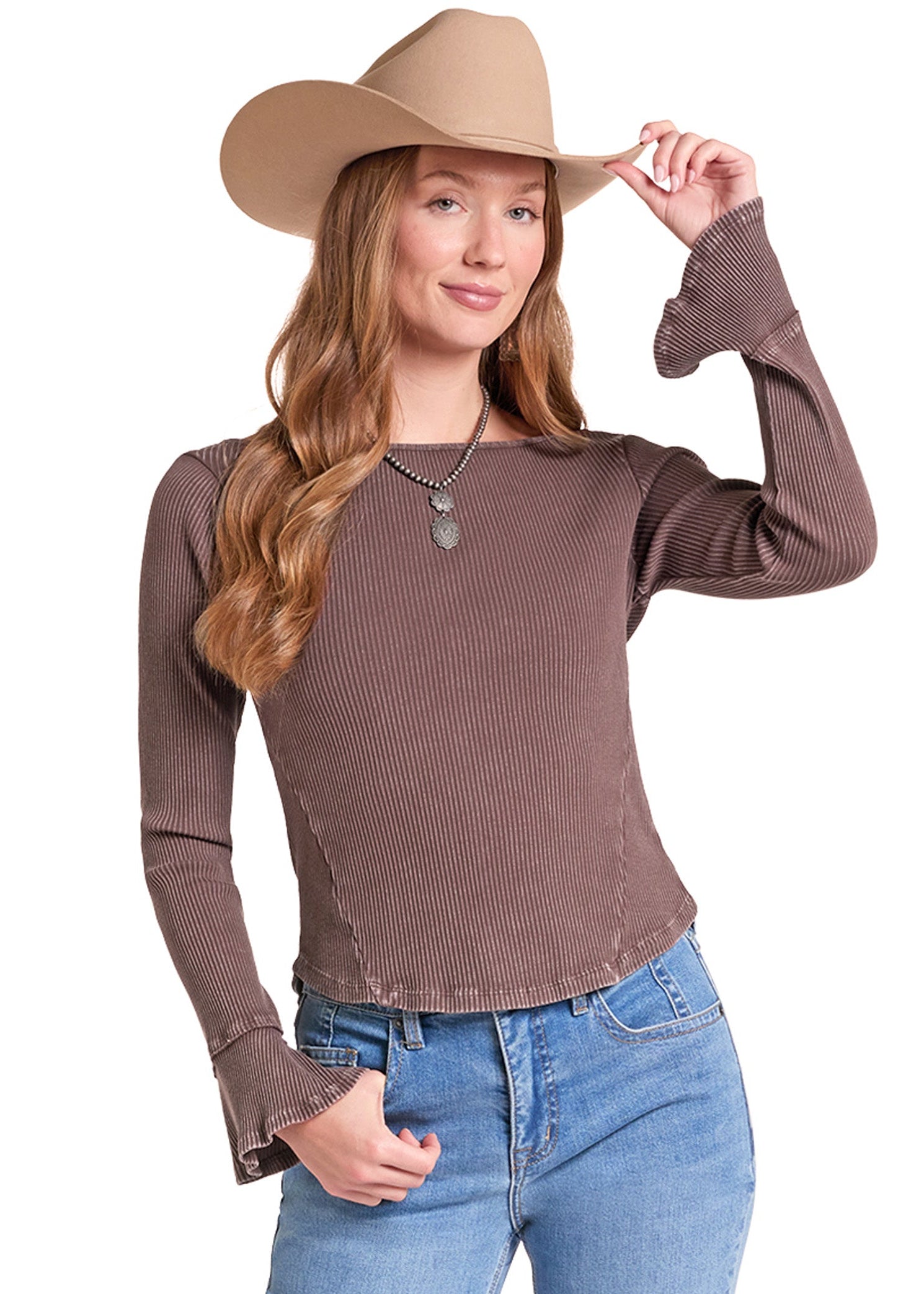 A woman wearing a brown ribbed long-sleeve top with flared cuffs, blue jeans, and a tan cowboy hat, posed against a plain white background.