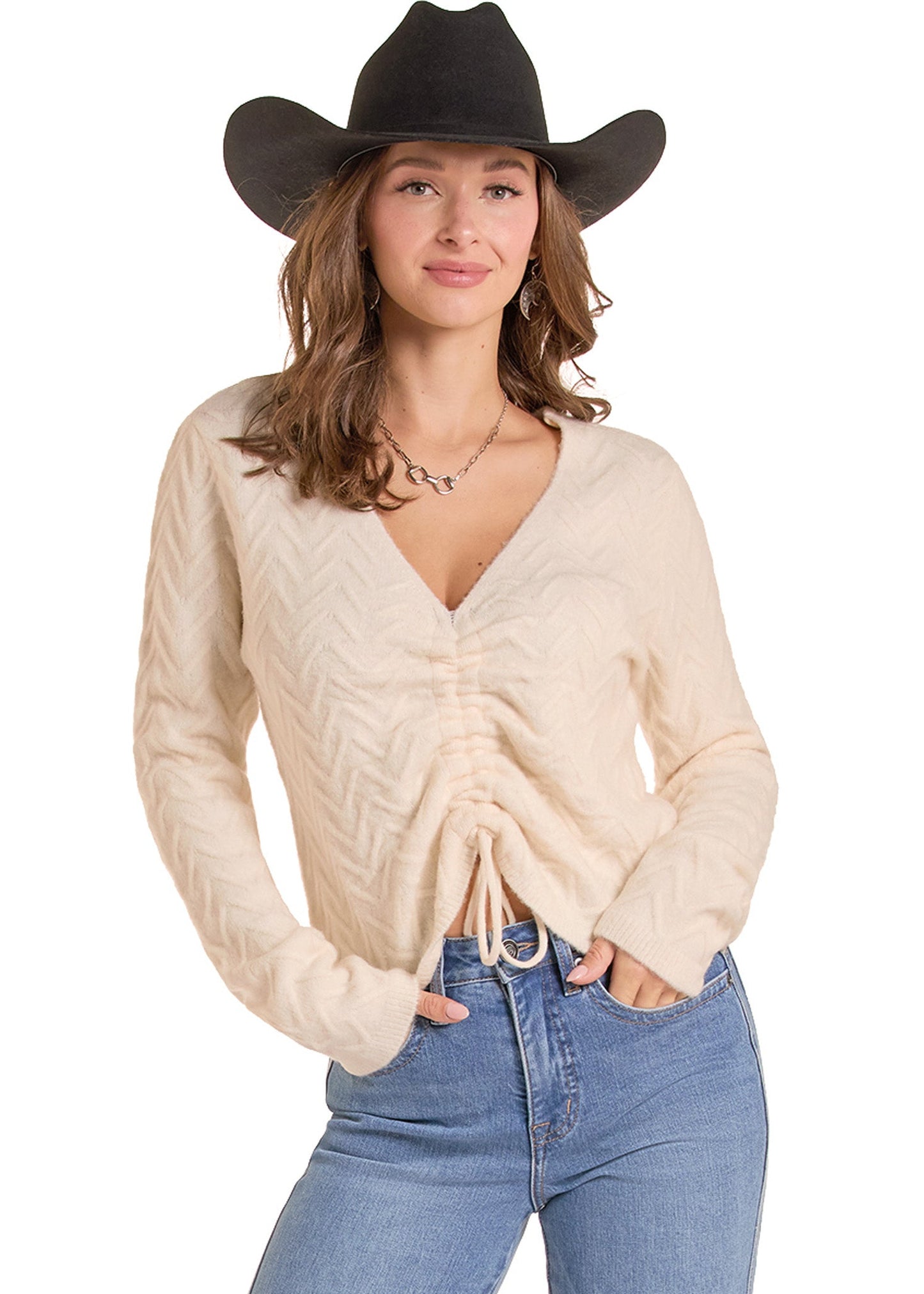 A woman wearing a cream-colored chevron knit sweater with a ruched front and drawstring hem, paired with blue jeans and a black cowboy hat, against a plain white background.