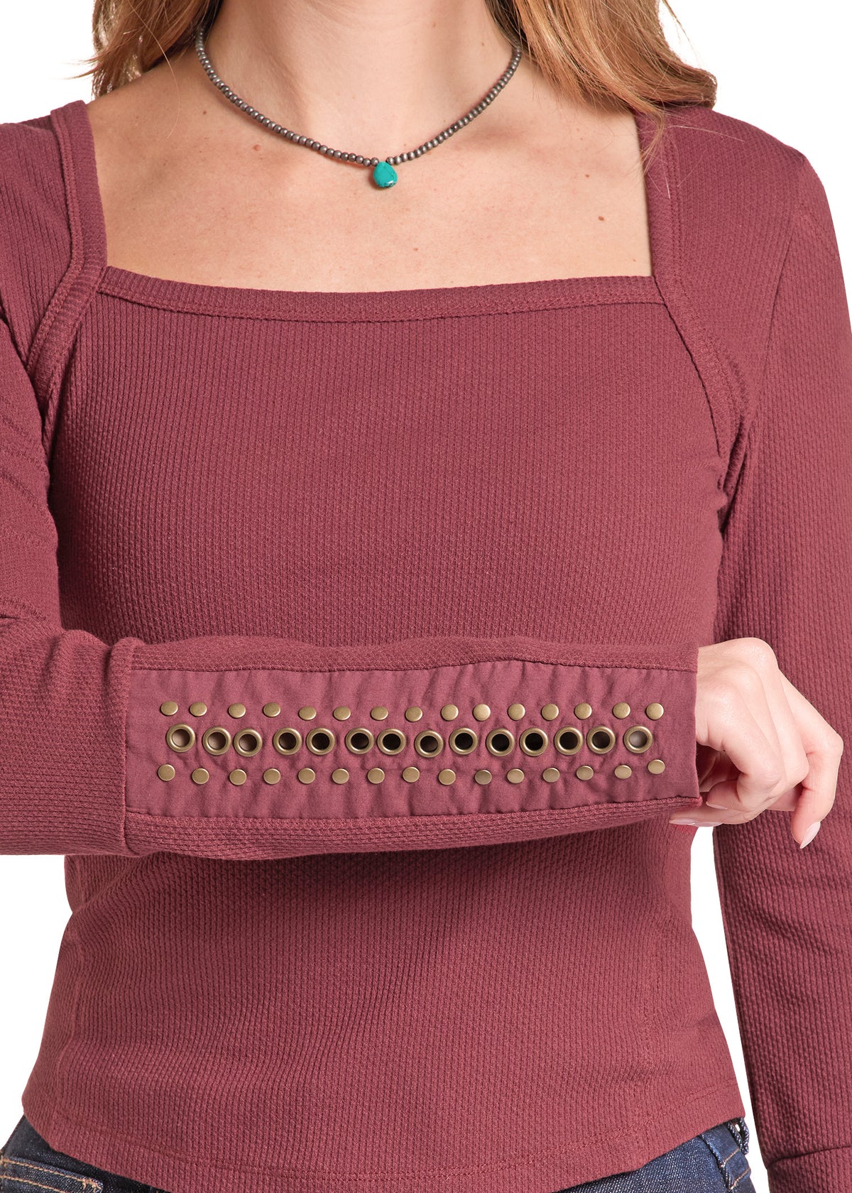 A woman wearing a ribbed long-sleeve top in dusty rose with a row of metallic eyelet embellishments across the upper sleeve cuff, shown against a plain white background.
