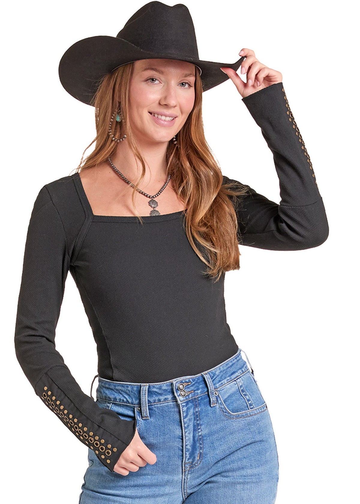 A woman wearing a black long-sleeve fitted top with decorative gold-tone stud detailing on the cuffs, paired with blue jeans and a black cowboy hat, against a plain white background.