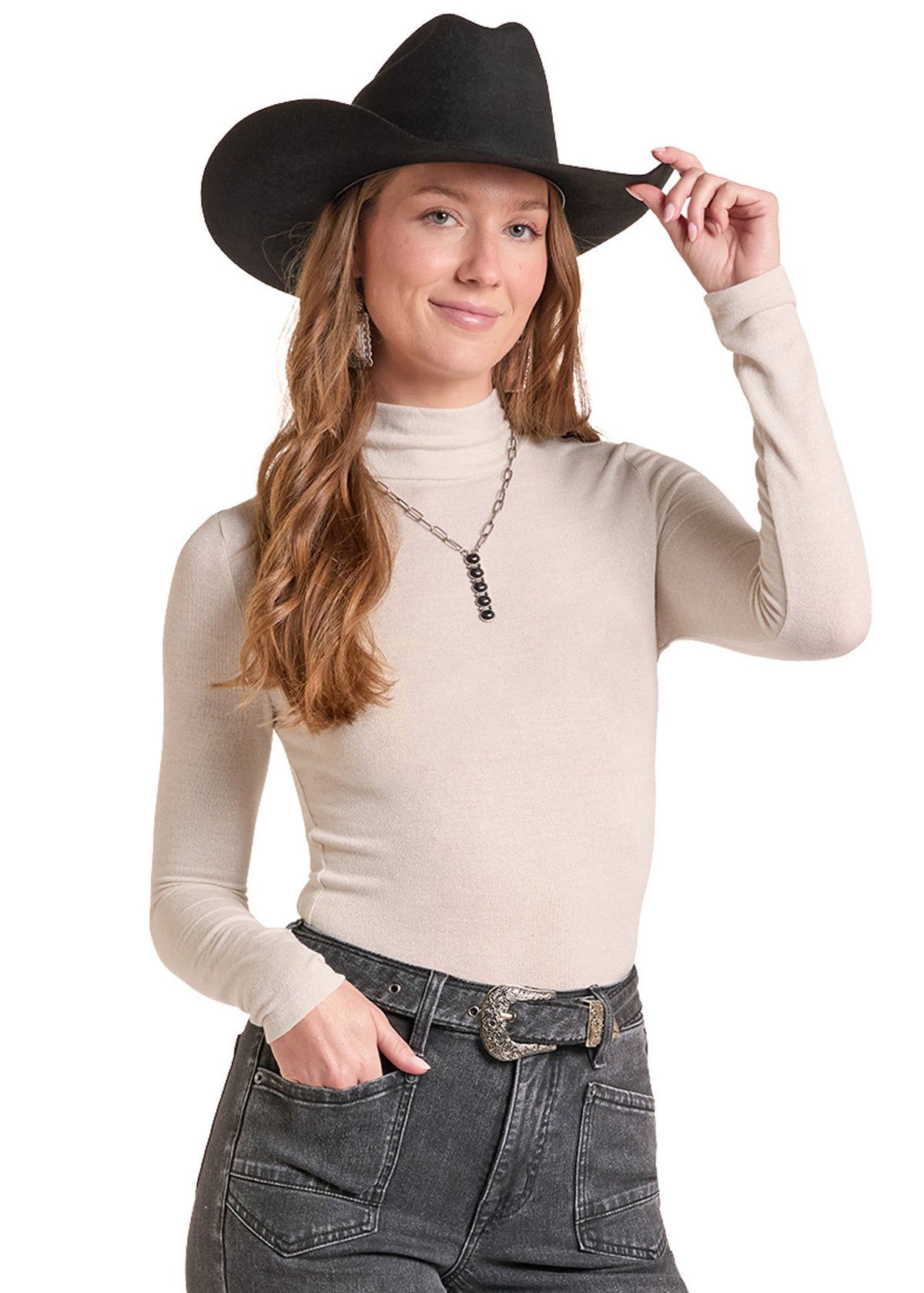 A woman wearing a black cowboy hat, a beige ribbed turtleneck sweater, and dark gray jeans, posing against a solid white background.