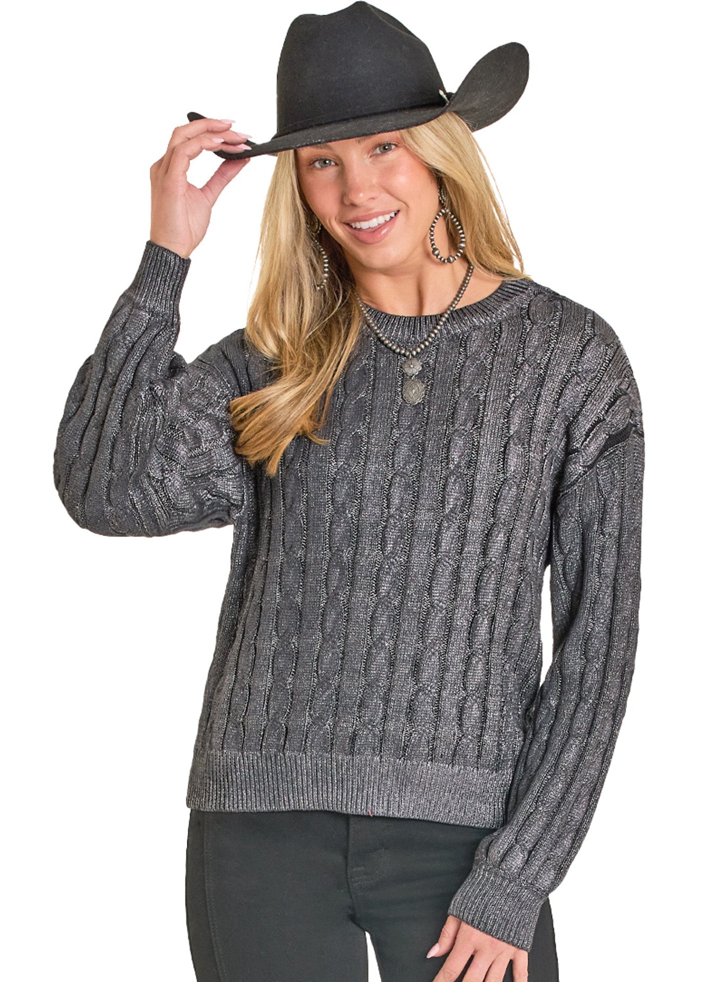 A smiling woman poses against a white background, holding her black cowboy hat while wearing the Rock & Roll Denim Foil Cable Knit Sweater in gray, paired with black pants, large hoop earrings, and a necklace.
