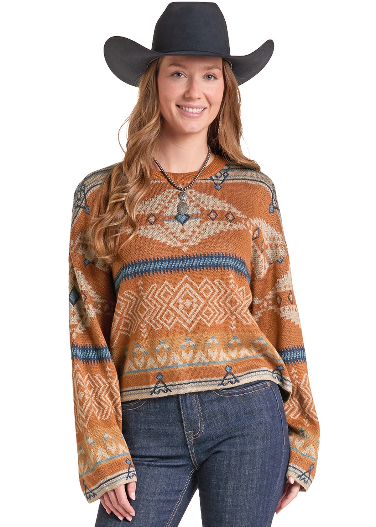 A woman wearing a rust-orange knit sweater with a Southwestern geometric pattern in beige, blue, and white, paired with a black cowboy hat and blue jeans, against a solid white background.