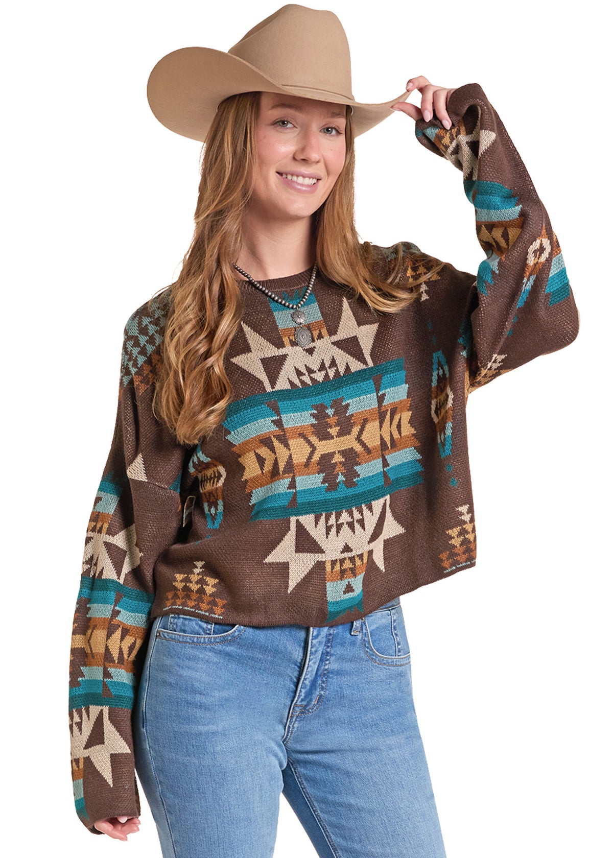 A woman wearing a brown long-sleeve knit sweater with a colorful Southwestern geometric pattern, paired with light blue jeans and a tan cowboy hat.