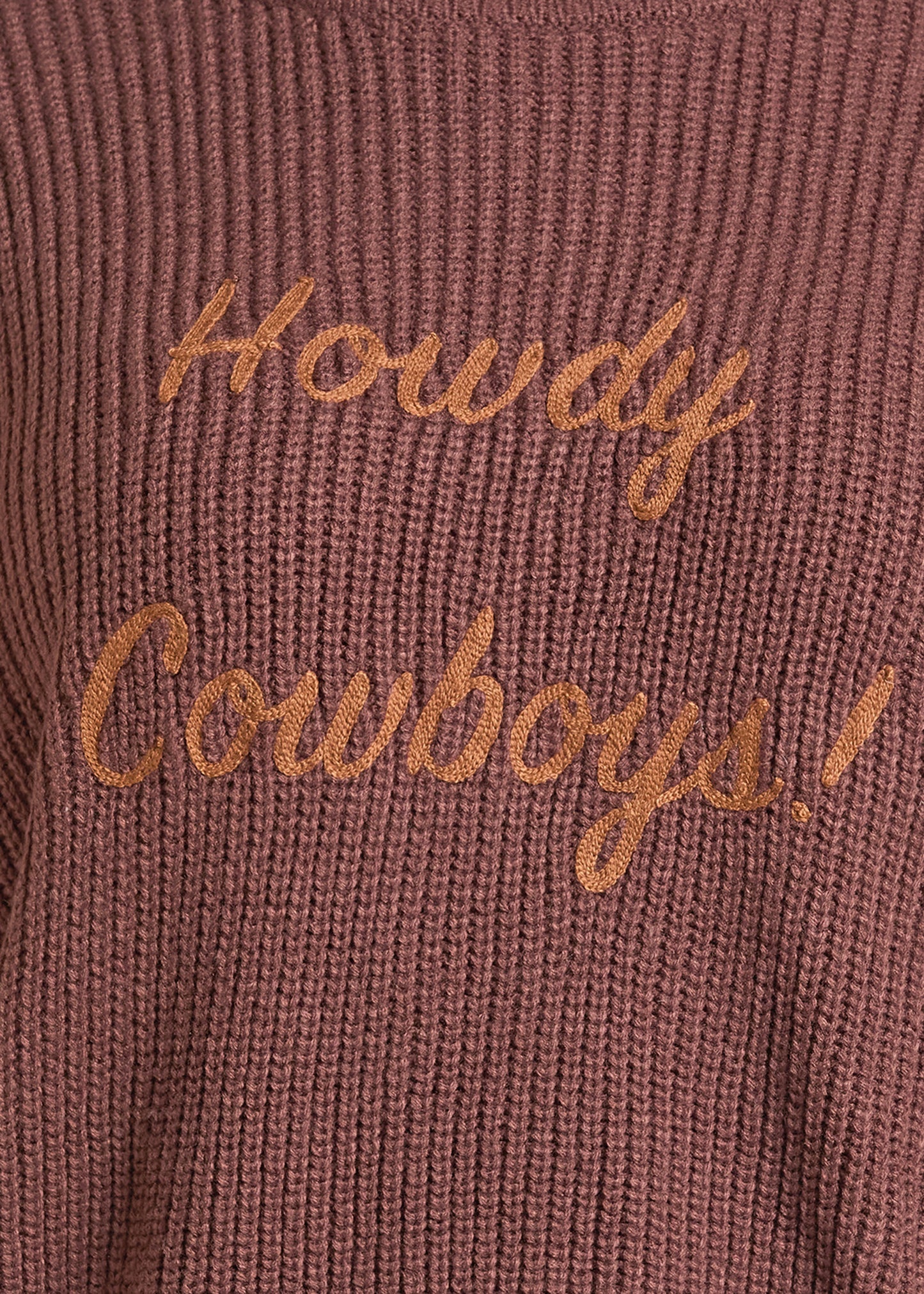 Close-up of a ribbed knit sweater in a dusty rose or mauve-brown color with embroidered cursive text reading 'Howdy Cowboys!' in a contrasting tan-gold thread.