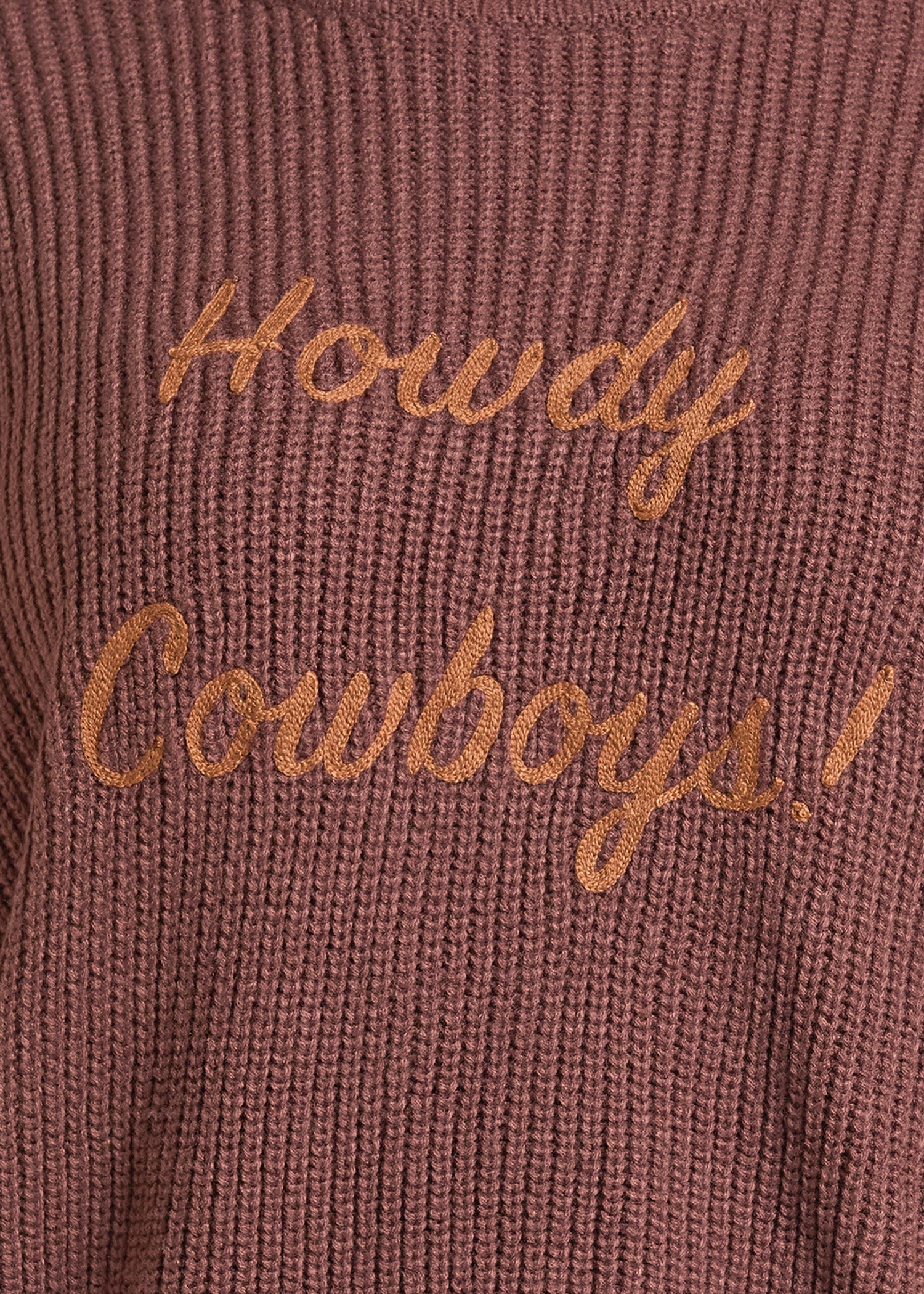 Close-up of a ribbed knit sweater in a dusty rose or mauve-brown color with embroidered cursive text reading 'Howdy Cowboys!' in a contrasting tan-gold thread.