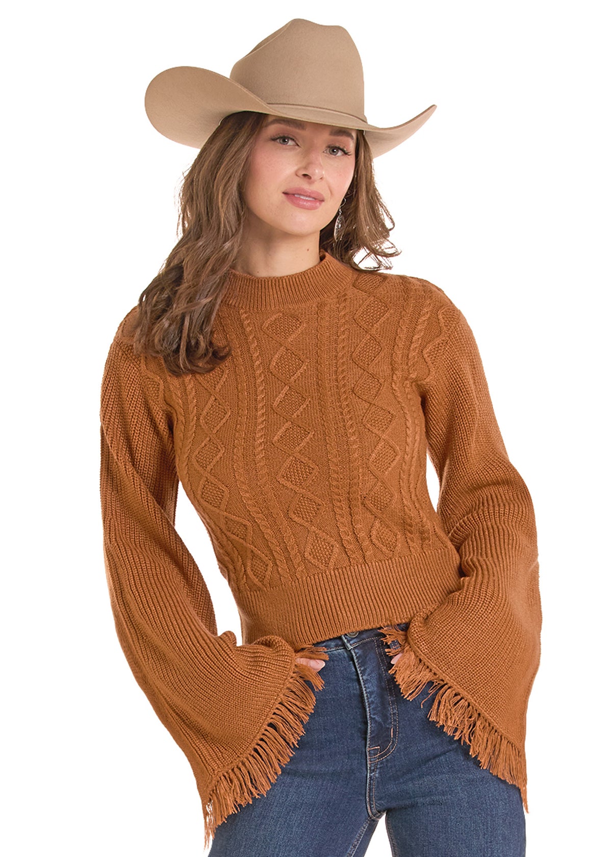 A woman wearing a cropped, long-sleeve knit sweater in a warm camel brown color with fringe trim, paired with blue jeans and a tan cowboy hat, against a solid white background.