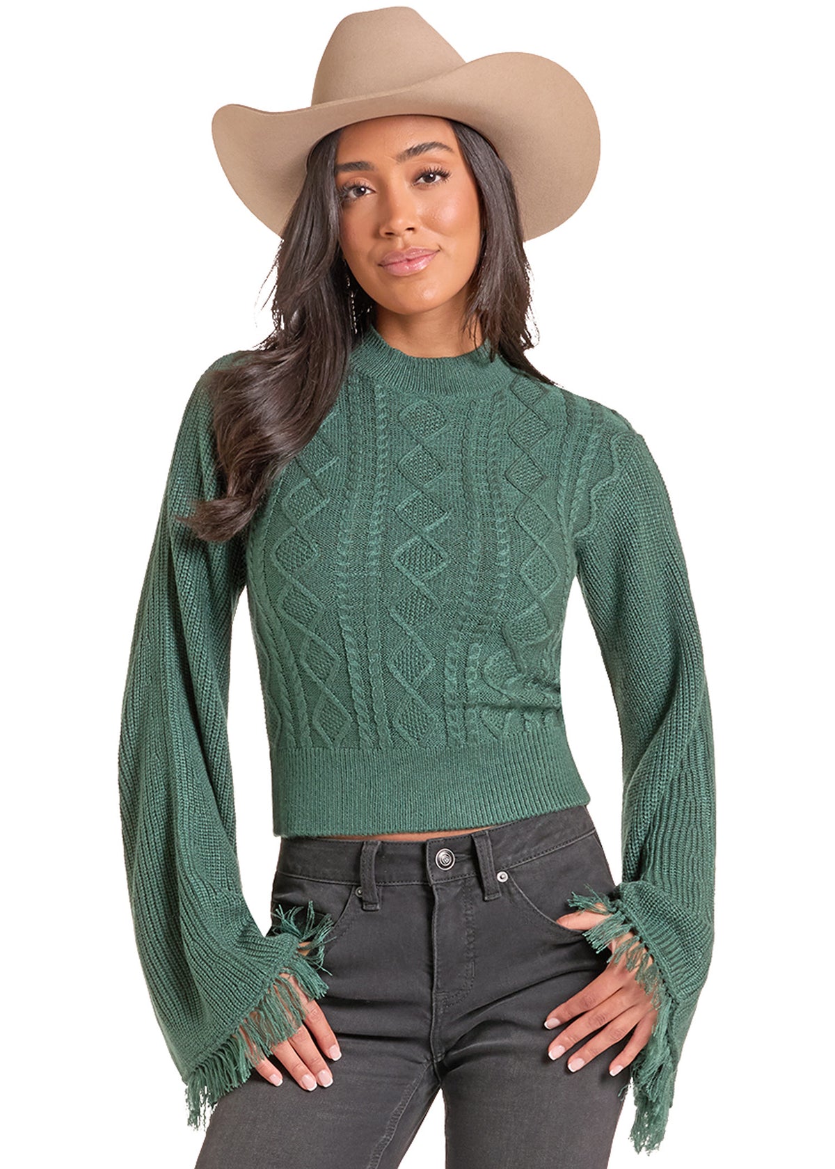 A woman modeling a green cable-knit cropped sweater with distressed fringed bell sleeves, paired with dark gray jeans and a beige cowboy hat, against a solid white background.