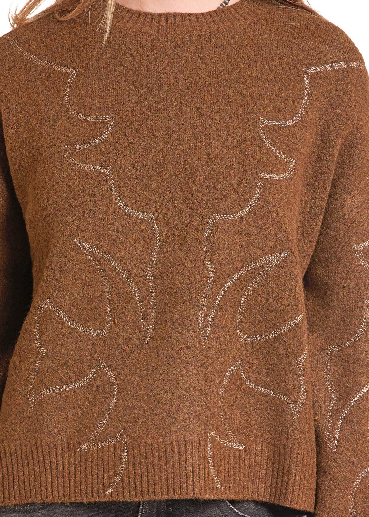 A close-up of a person wearing a brown knit sweater with a subtle tonal leaf pattern and dark gray jeans against a plain white background.