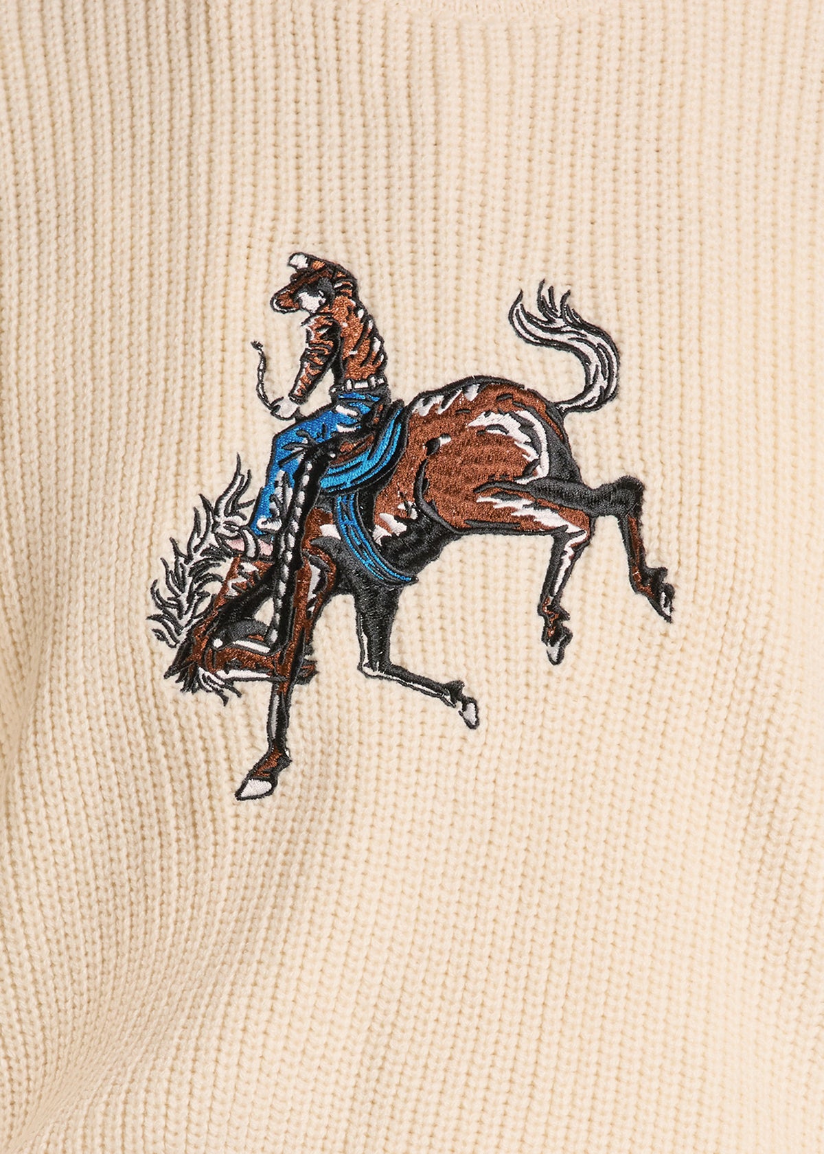 Close-up of a cream-colored ribbed knit fabric featuring an embroidered graphic of a cowboy riding a bucking brown horse with a blue saddle.