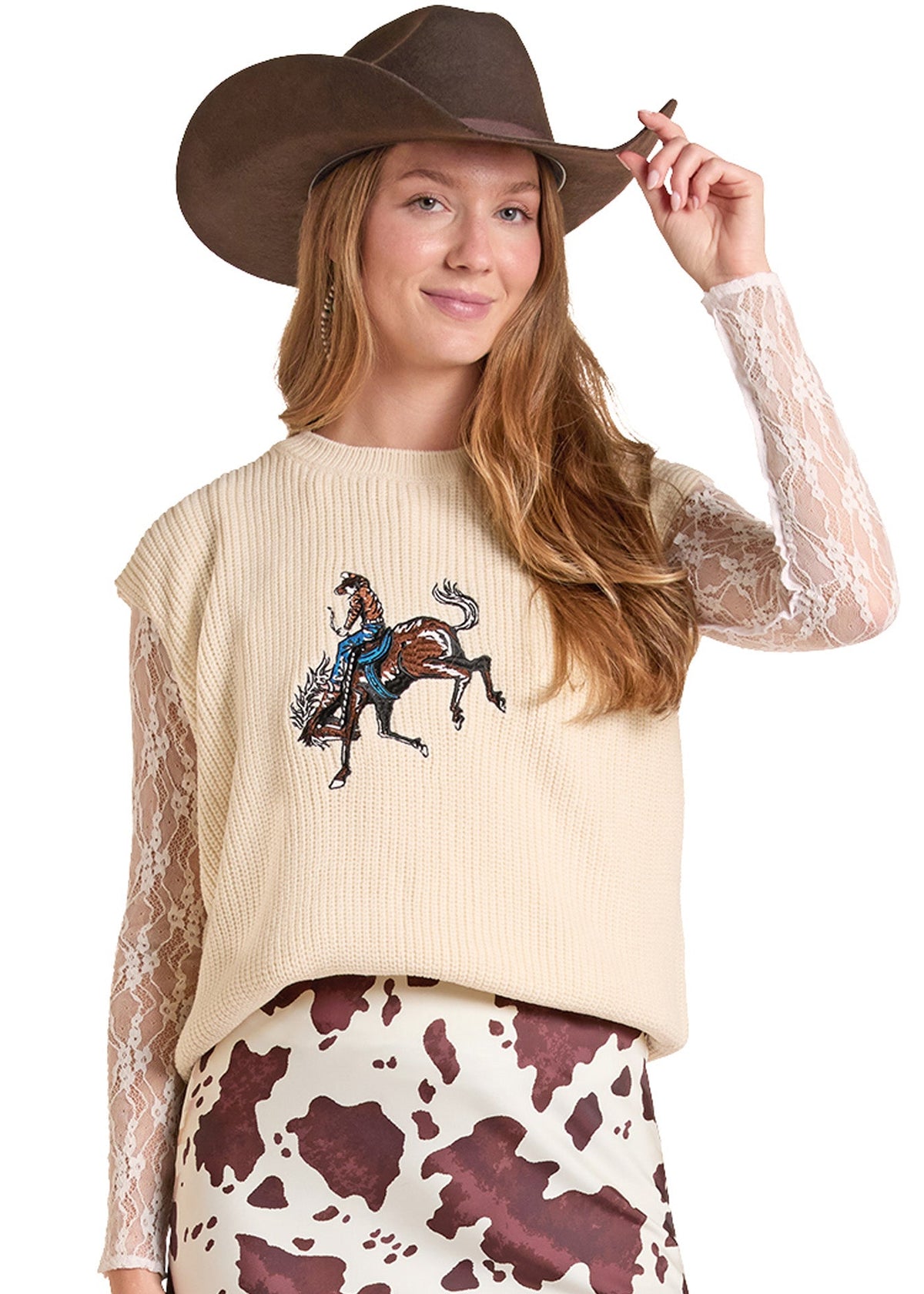 A woman wearing a cream ribbed knit cropped sweater vest with a western rider graphic, sheer lace long sleeves, a brown cowboy hat, and a brown and white cow print skirt, set against a plain white background.