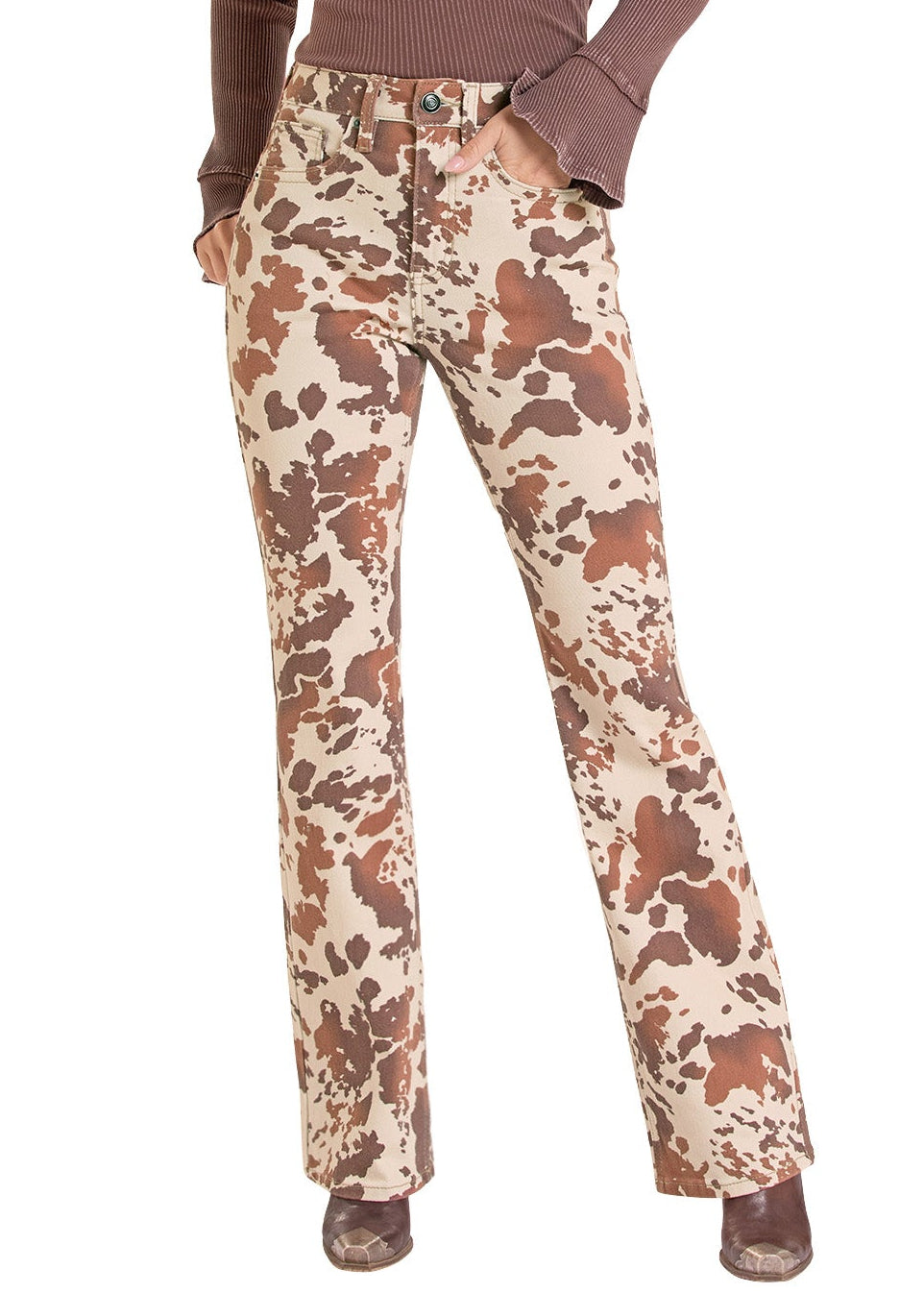 A woman modeling a western-style outfit consisting of a brown long-sleeve top, high-waisted cream and brown cow-print pants, and a tan cowboy hat, set against a plain white background.