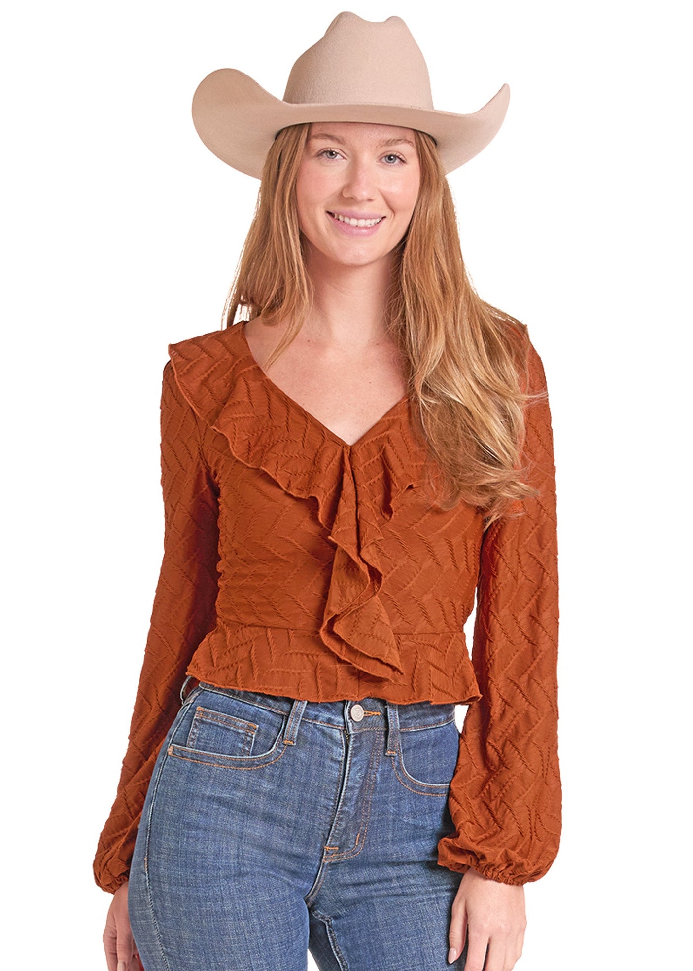 A woman wearing a rust-orange long-sleeve ruffled blouse, blue jeans, and a beige cowboy hat, photographed against a plain white background.