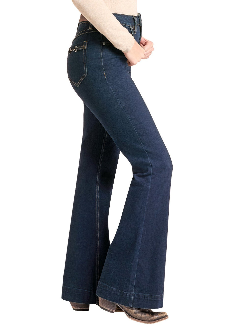 A woman modeling dark blue flare jeans, a beige long-sleeve top, and a black cowboy hat, shown in profile against a solid white background.