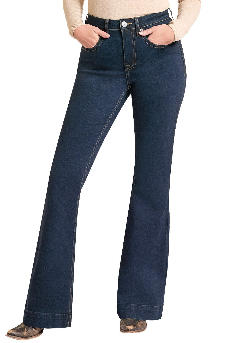 A woman modeling dark blue flare jeans with a beige long-sleeve top and a black cowboy hat, standing against a plain white background.