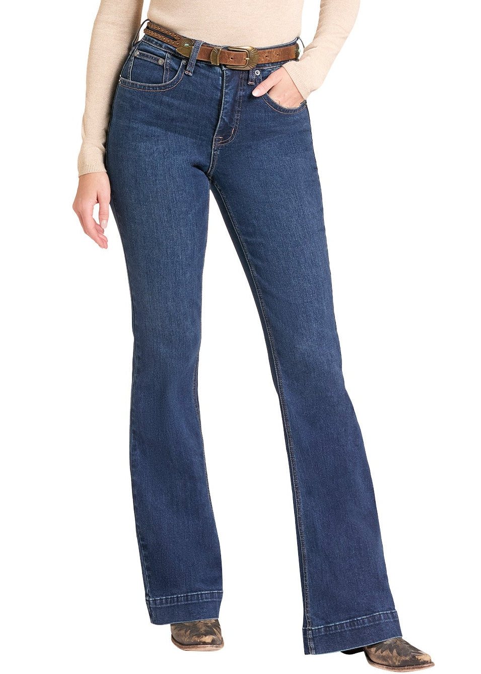 A woman modeling blue flare jeans with a brown belt, paired with a beige long-sleeve top, brown cowboy boots, and a beige cowboy hat, set against a plain white background.