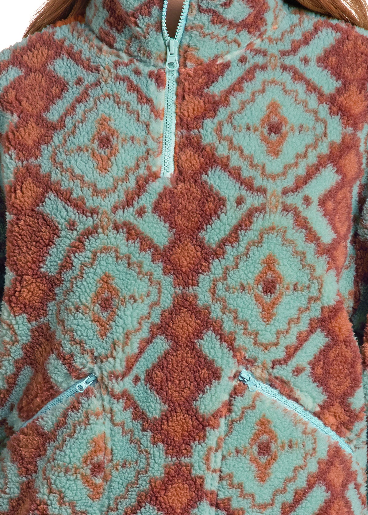 Close-up of a fleece quarter-zip pullover with a geometric diamond pattern in sage green and rust orange, featuring a light blue zipper.
