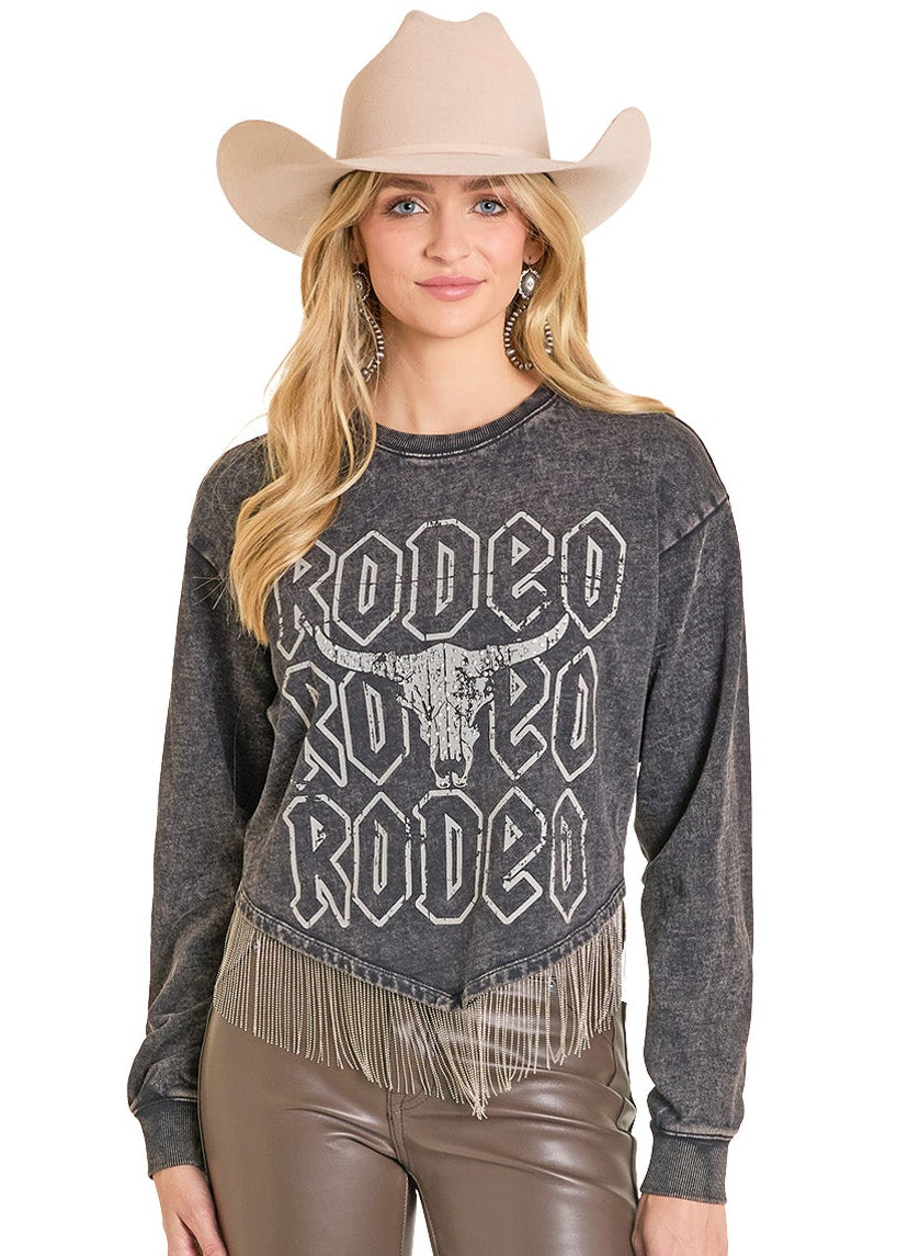 A woman wearing a light cowboy hat, earrings, brown leather pants, and the Rock & Roll Denim Chain Fringe Rodeo Graphic Pullover with a bull skull design stands facing forward and smiling.