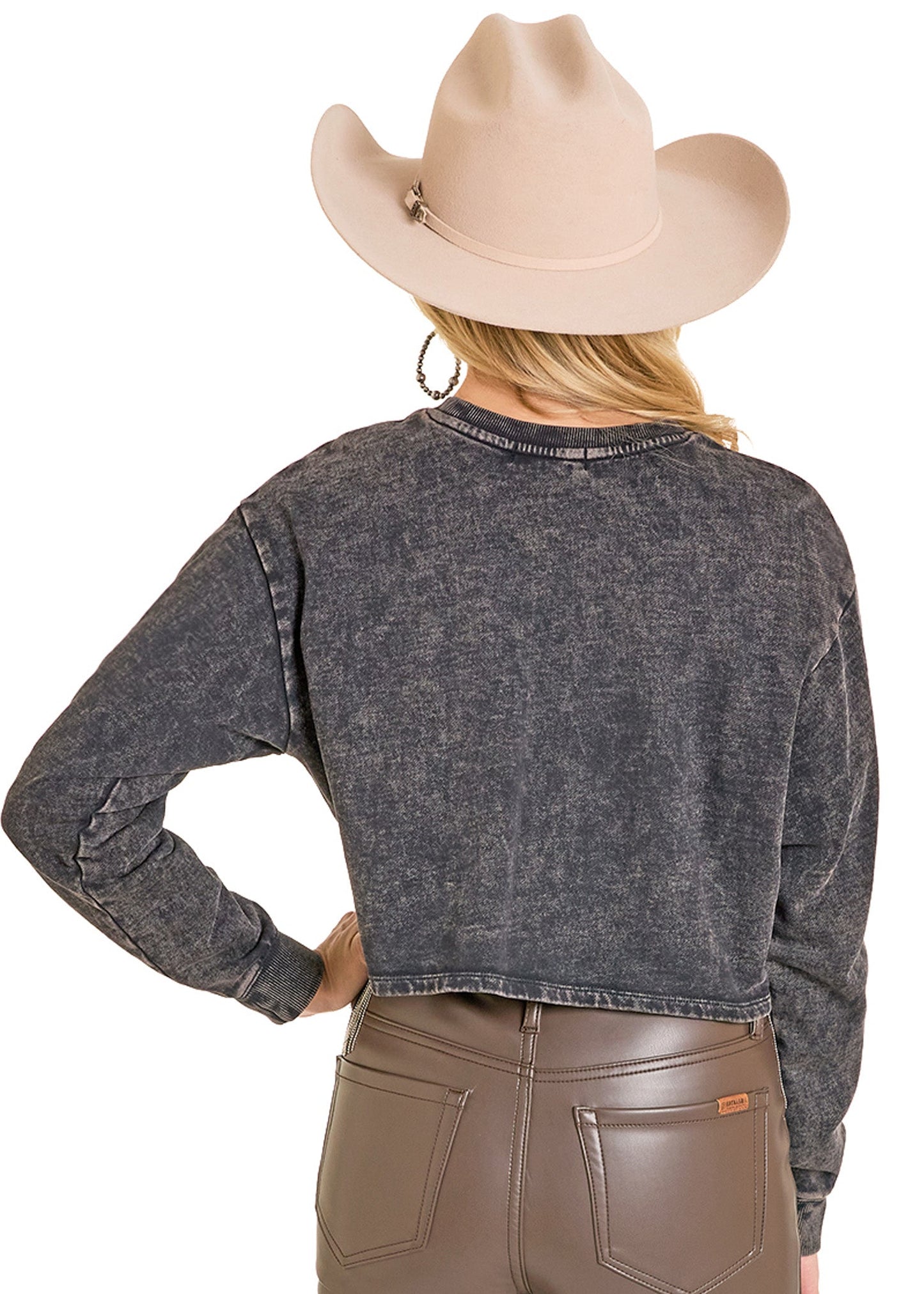 A person shown from the back wears a beige cowboy hat, the Rock & Roll Denim Chain Fringe Rodeo Graphic Pullover in faded black, and high-waisted brown leather pants.