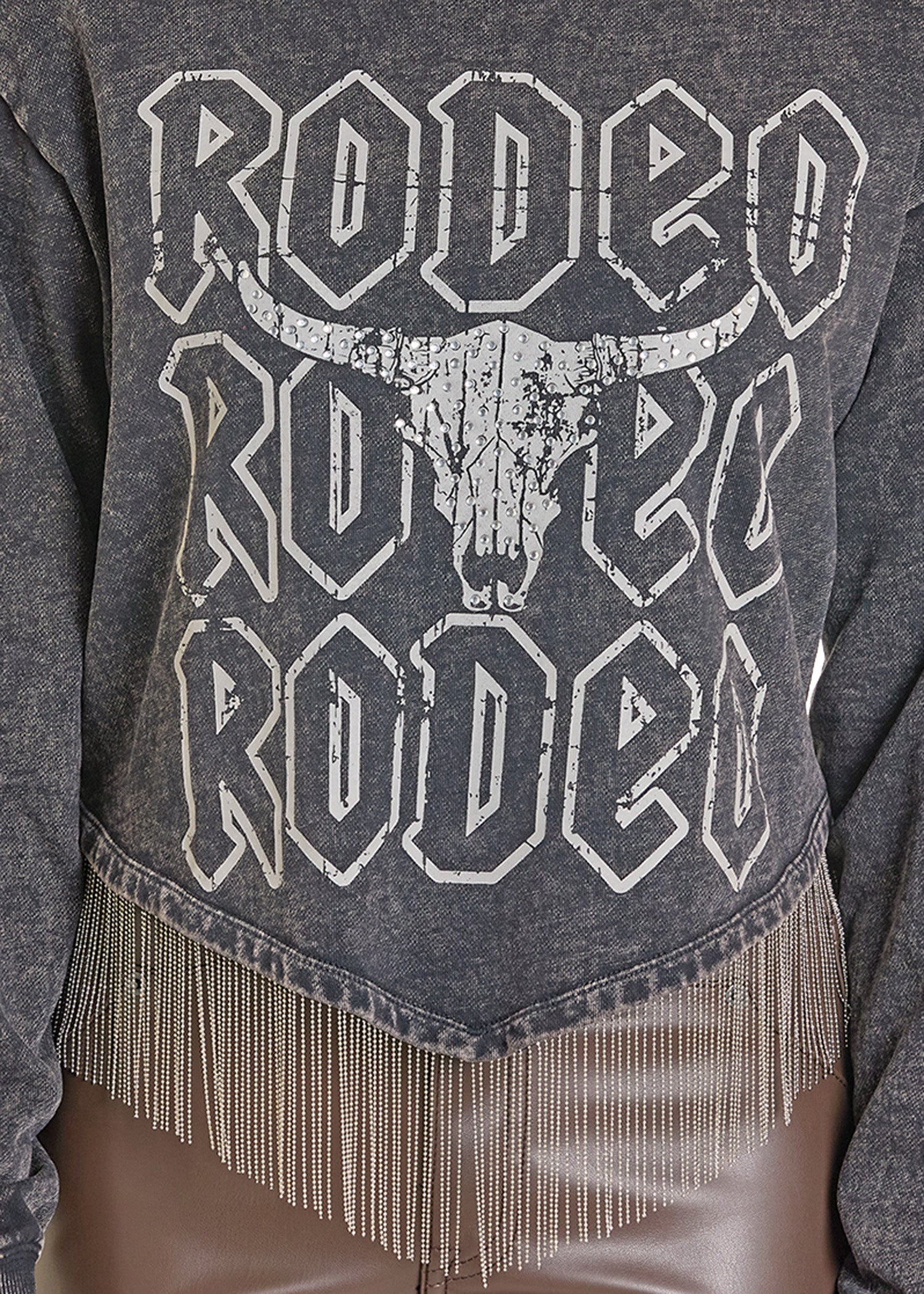 The Rock & Roll Denim Chain Fringe Rodeo Graphic Pullover features bold, distressed white “RODEO” lettering and a bull skull on black fabric, finished with silver chain fringe at the hem for an edgy statement look.