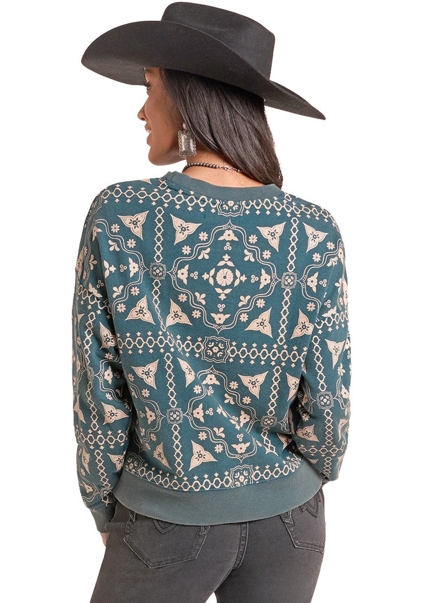 A woman shown from the back wearing a teal and beige patterned knit sweater, dark gray jeans, and a black cowboy hat against a plain white background.