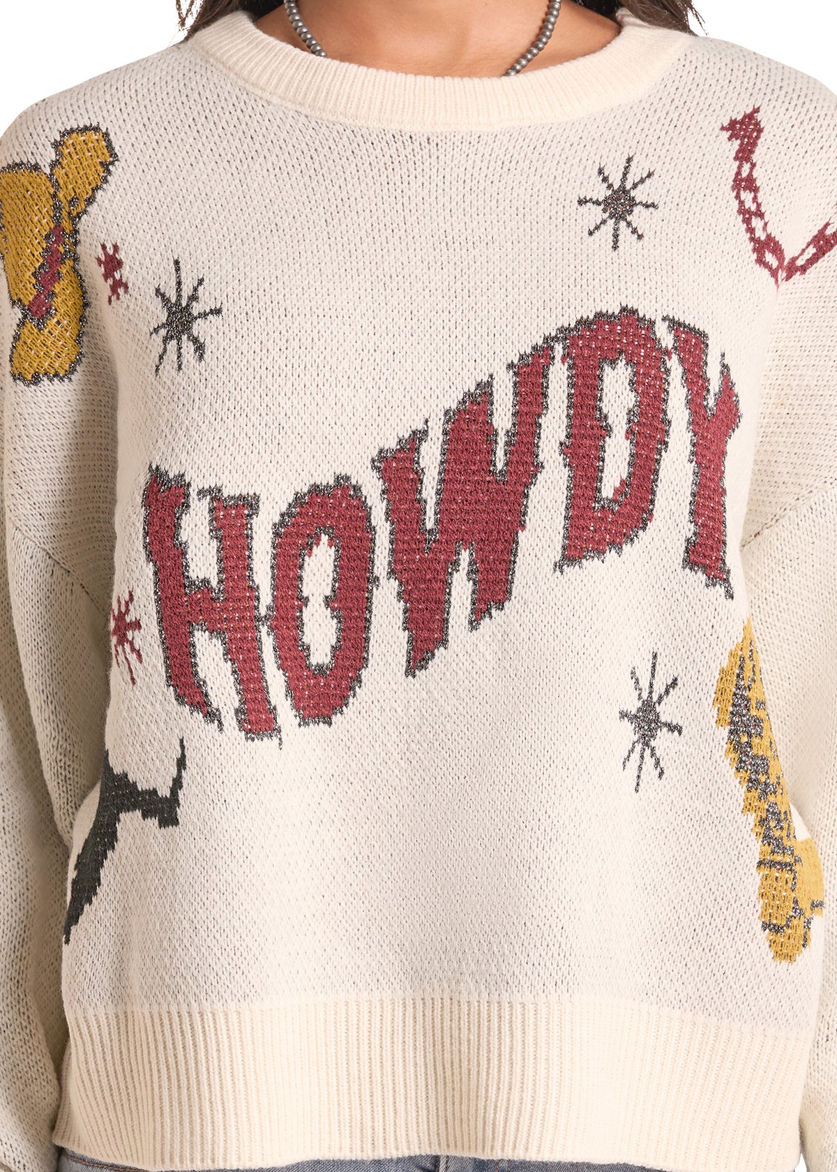 A close-up of a cream-colored knit sweater featuring the word 'HOWDY' in large red letters across the chest, with yellow and red decorative motifs and black starburst details.