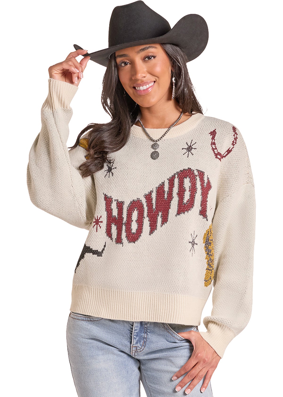 A woman wearing a cream-colored knit sweater with 'HOWDY' and western-themed embroidery, paired with a black cowboy hat and light blue jeans, posing against a white background.