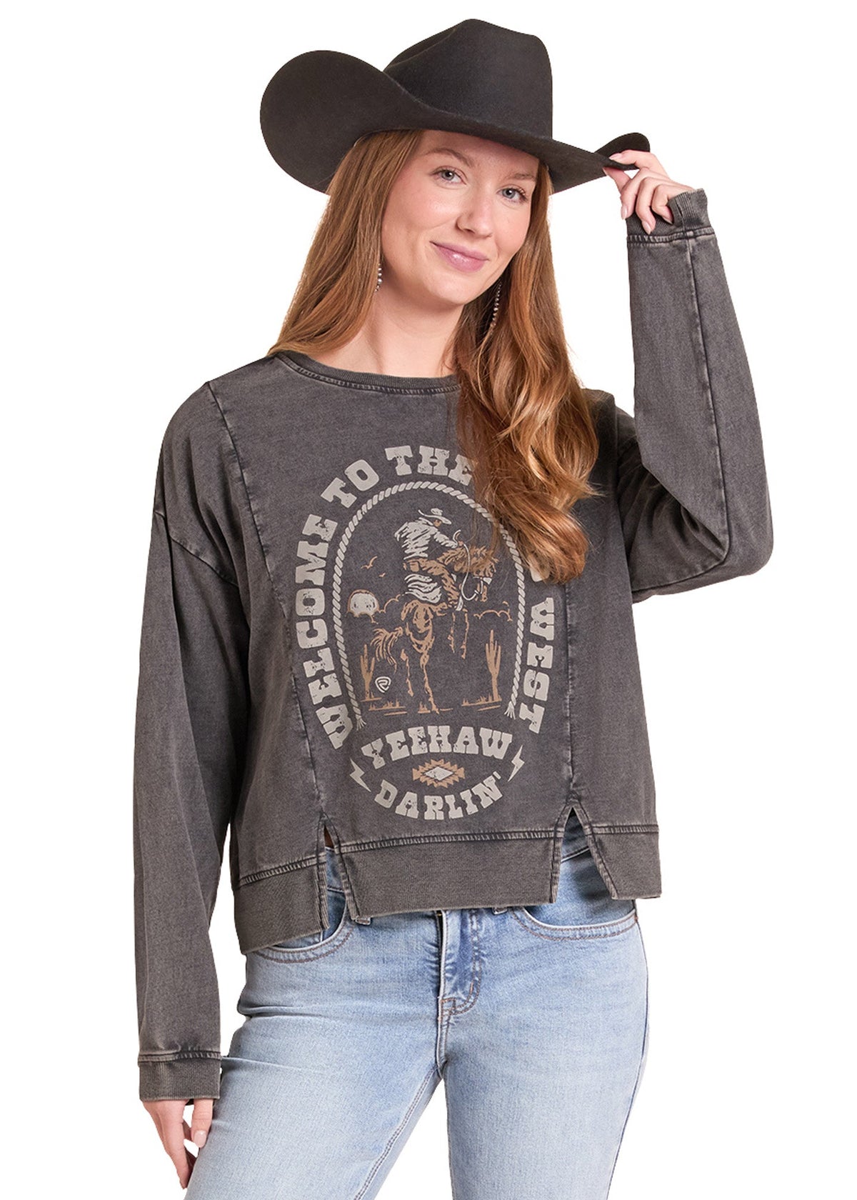 A woman wearing a faded charcoal gray graphic sweatshirt with a distressed western design, light blue jeans, and a black cowboy hat, standing against a white background.