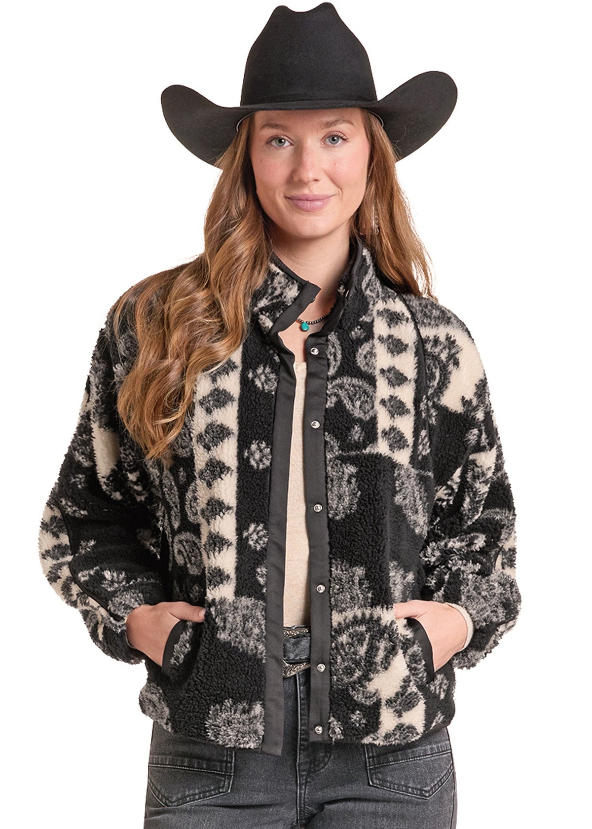 A woman wearing a black and beige patterned sherpa fleece button-up jacket, a black cowboy hat, and gray jeans, standing against a plain white background.