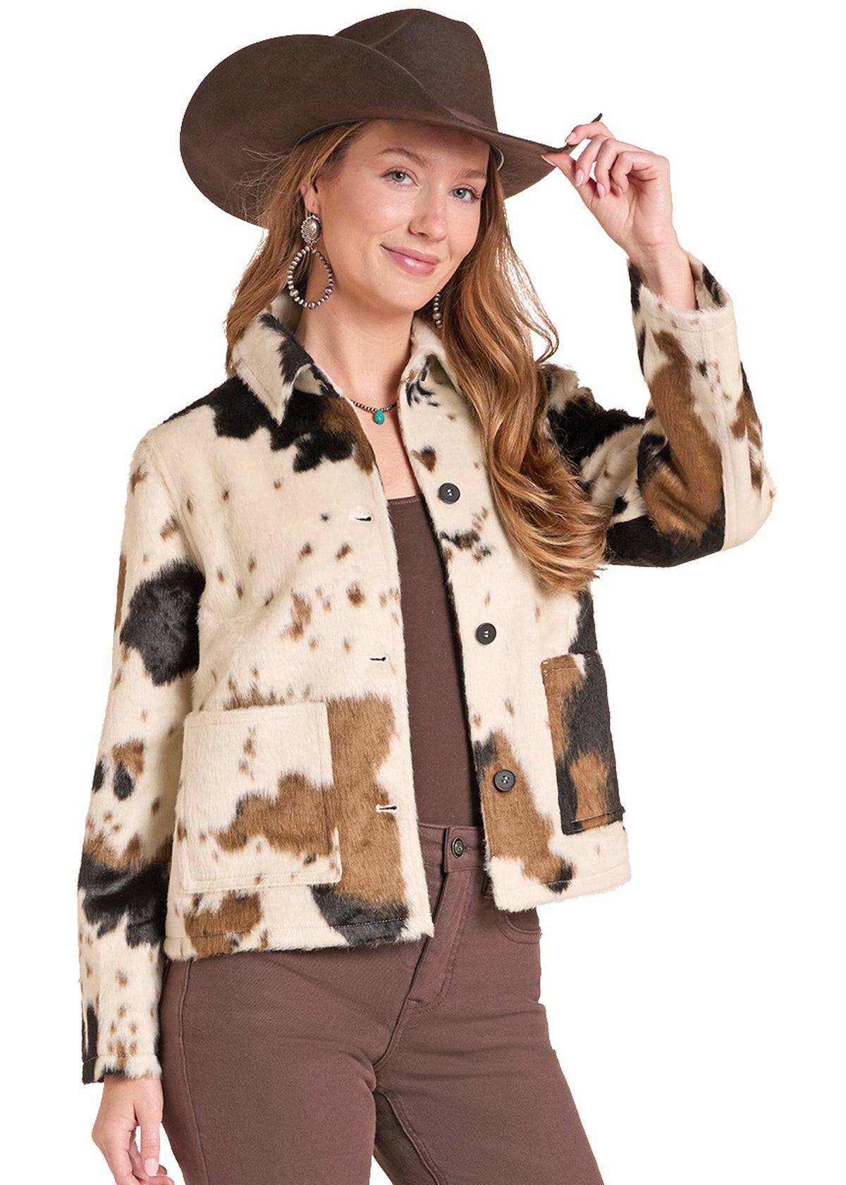 A woman wearing a brown cowboy hat and a cropped faux-fur jacket with a cow-print pattern in beige, brown, and black. She is also wearing brown pants and large silver hoop earrings, posing against a solid white background.