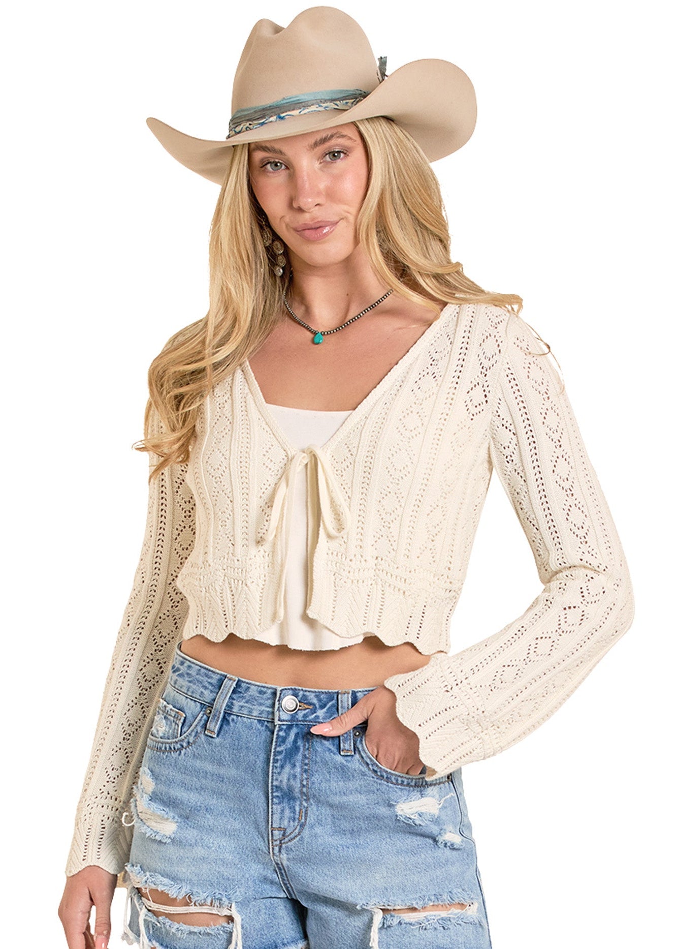 Woman wearing a beige lace cardigan, white top, and denim shorts with a cowboy hat on a white background