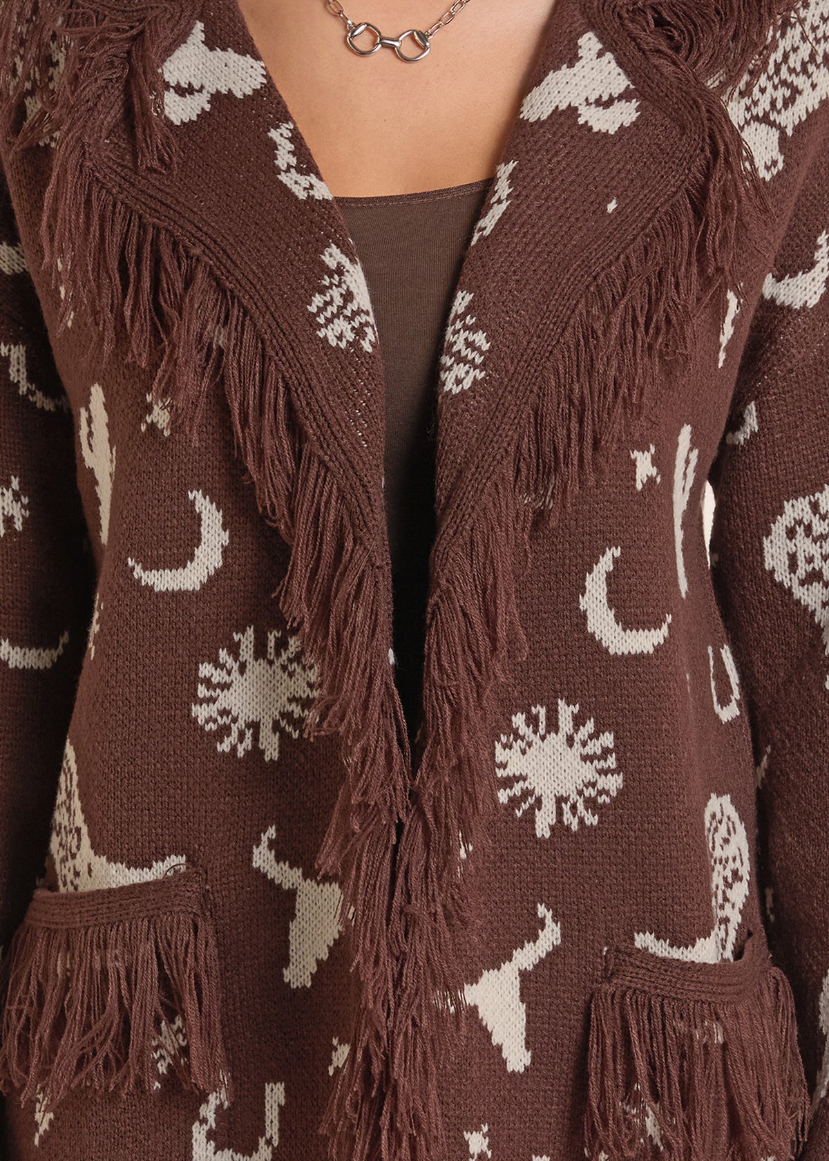 A close-up of a brown knit cardigan with cream-colored celestial patterns (moons, suns, stars) and long fringe trim, worn over a brown top.