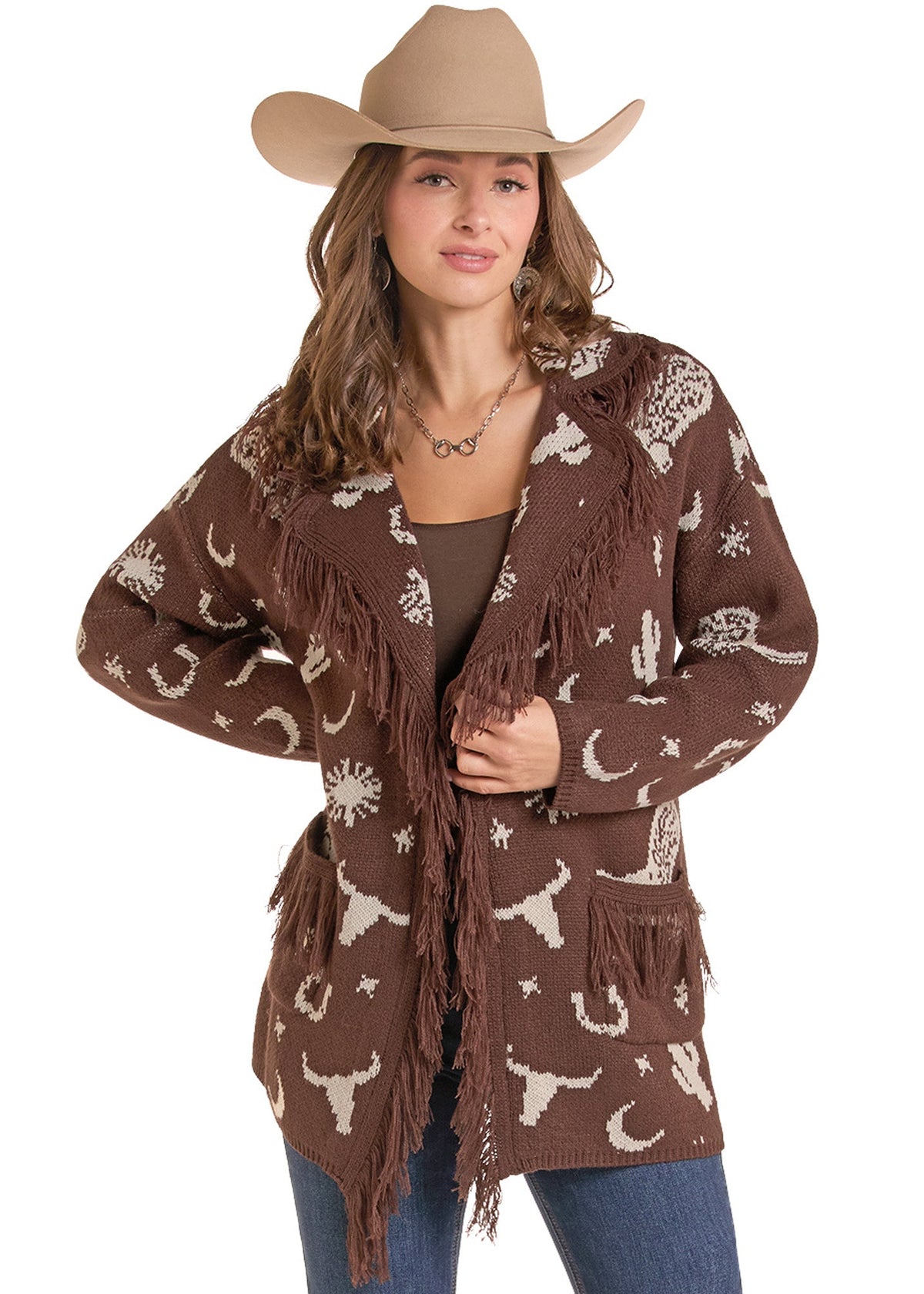 A woman modeling a brown open-front fringe cardigan with a white western print of stars, moons, and cacti, paired with a tan cowboy hat, brown top, and blue jeans against a white background.