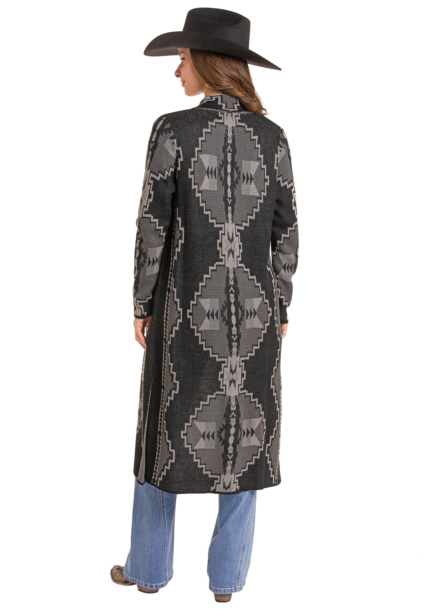 A woman shown from the back wearing a long dark gray and black patterned coat with geometric Southwestern-style motifs, blue jeans, and a black cowboy hat against a plain white background.