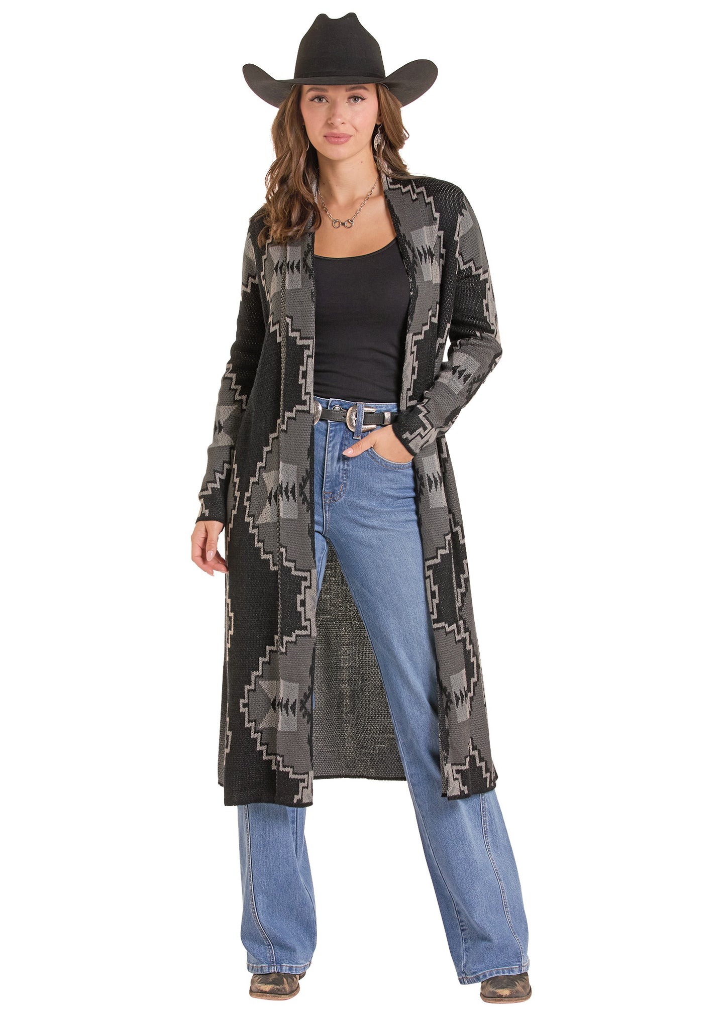 A woman modeling a long open-front cardigan with a black and gray geometric Southwestern pattern, styled with a black cowboy hat, black top, blue jeans, and brown boots against a white background.